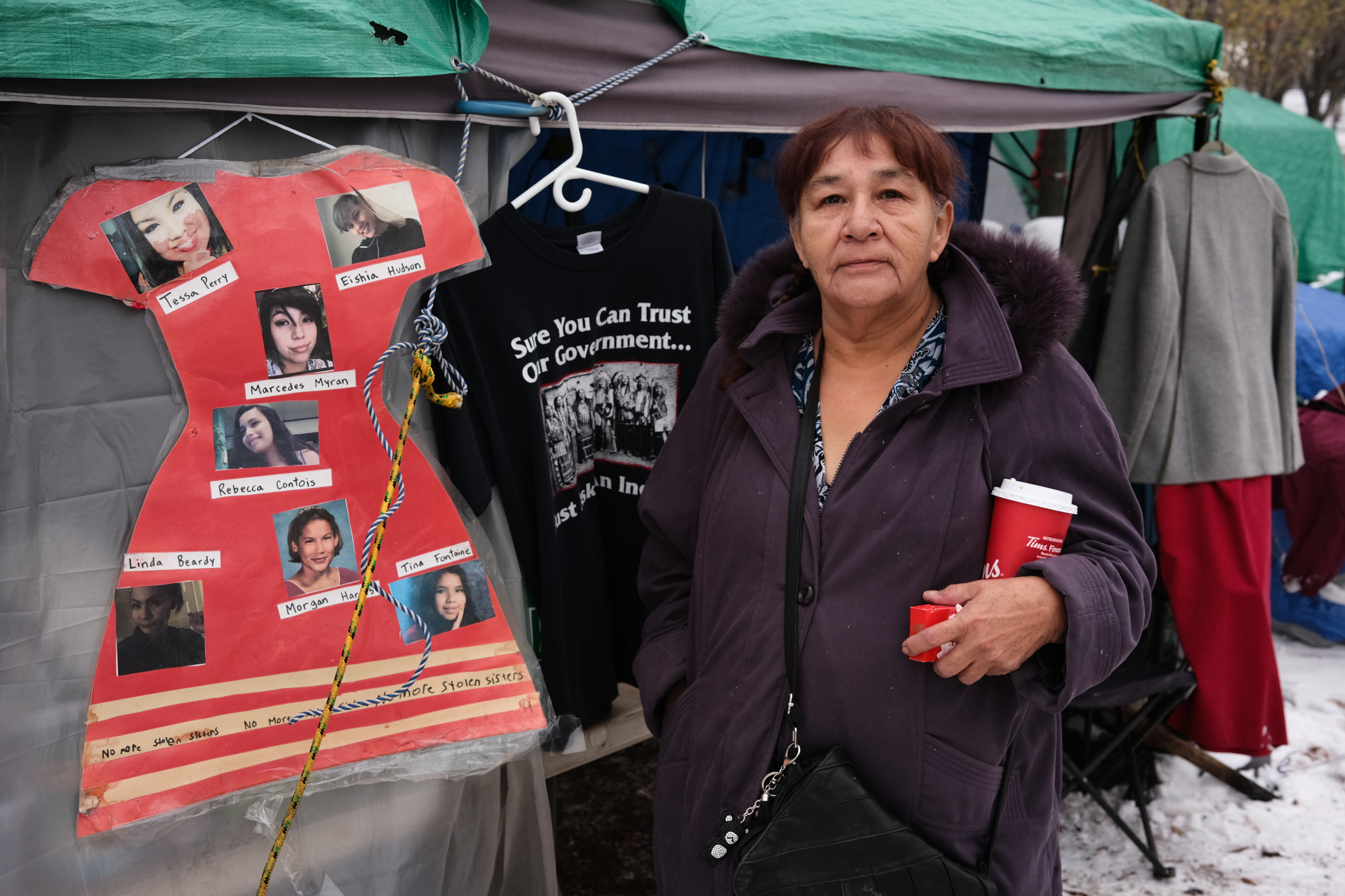 A woman in a black winter coat holds a red coffee cup in the crook of one of her arms, while her other hand is in her pocket. Snow is visible behind her, as is a protest camp. Pinned to a nearby structure is a dress cut from red paper, decorated with the photographs of missing Indigenous women.
