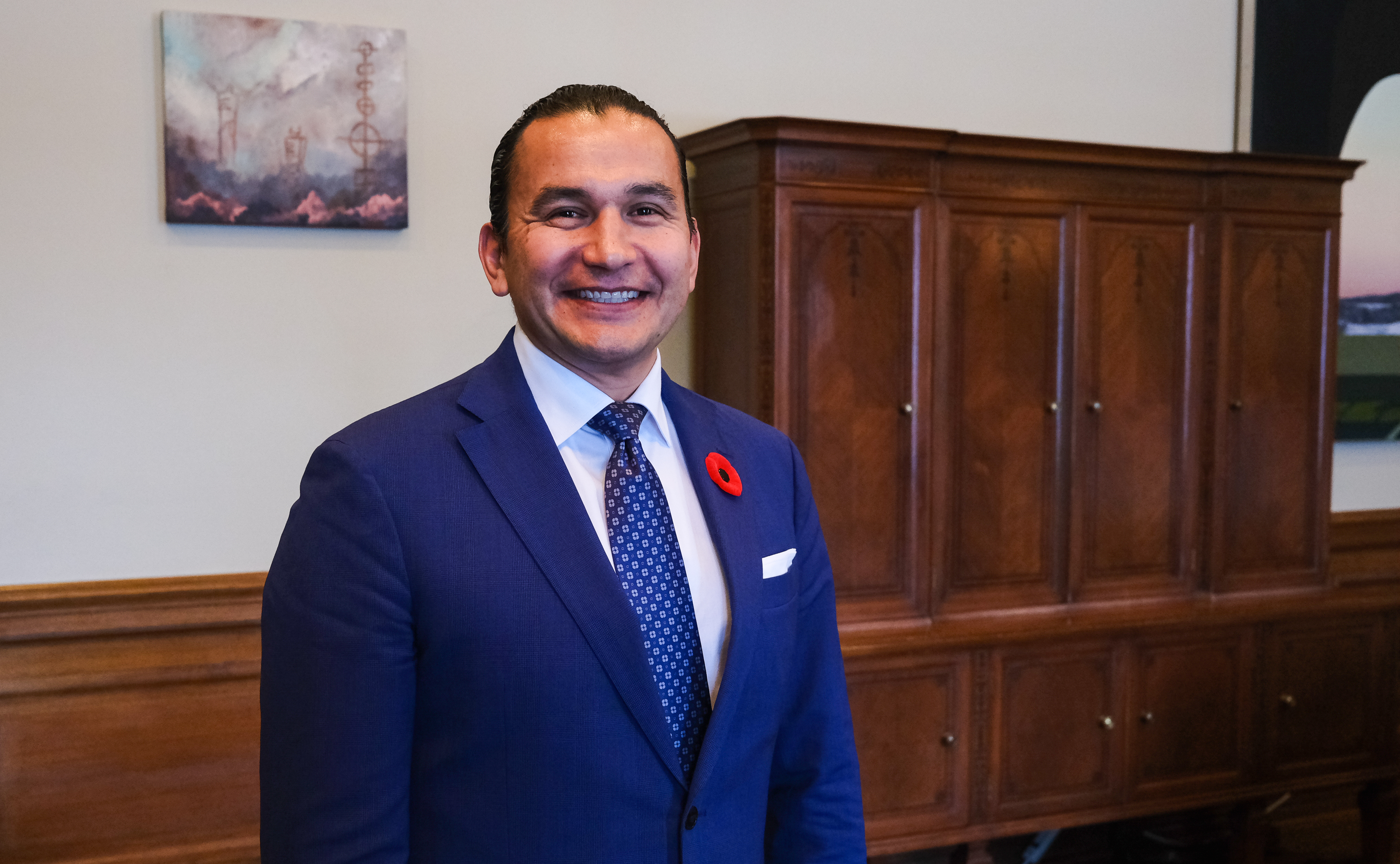 Wab Kinew, dressed in a blue suit and matching blue tie with a red poppy pin in the lapel, smiles as he faces the camera. A brown wardrobe is visible behind him.