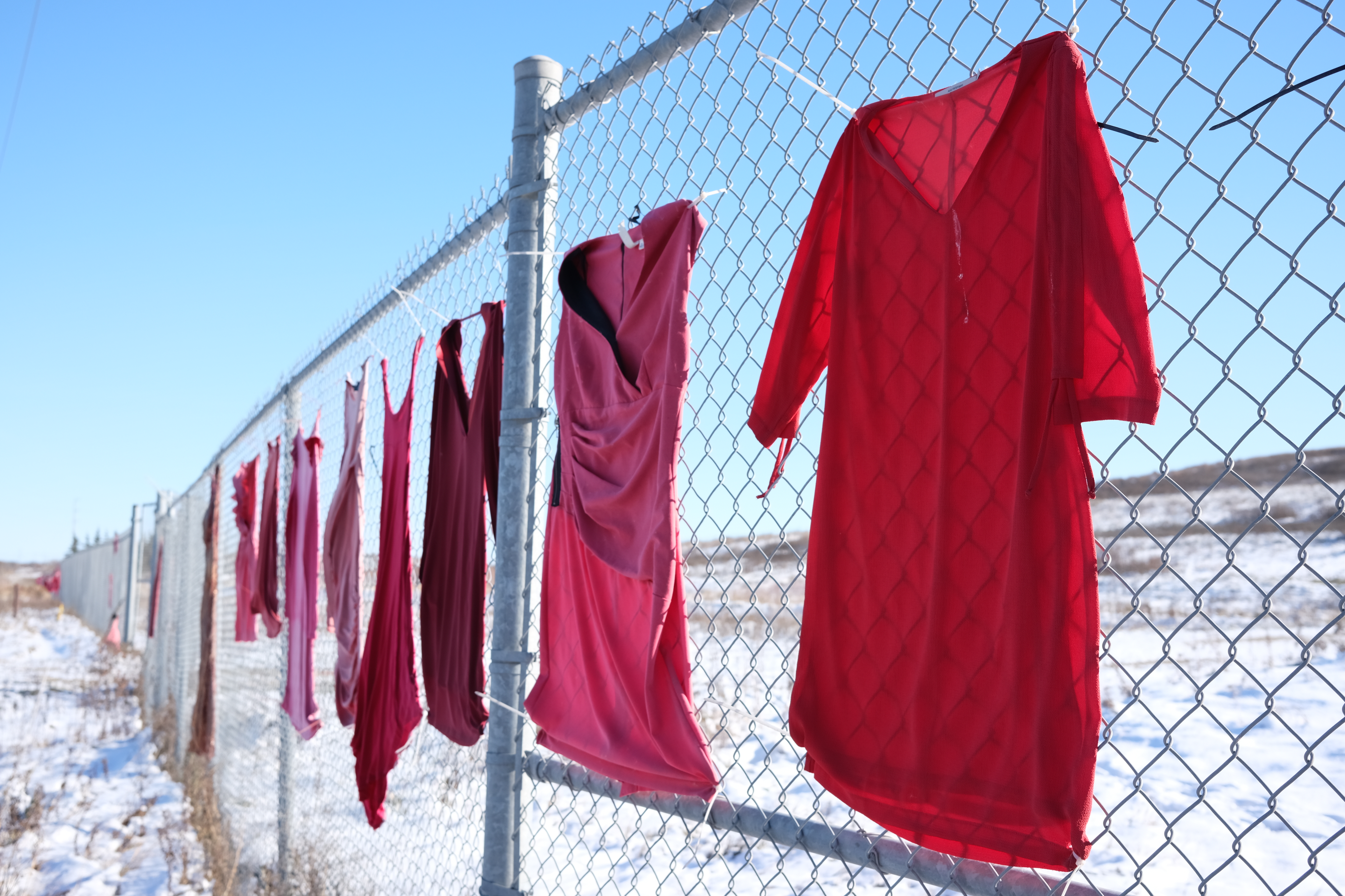 A row of red dresses are pinned on a chain-link fence, amid a snowy landscape.