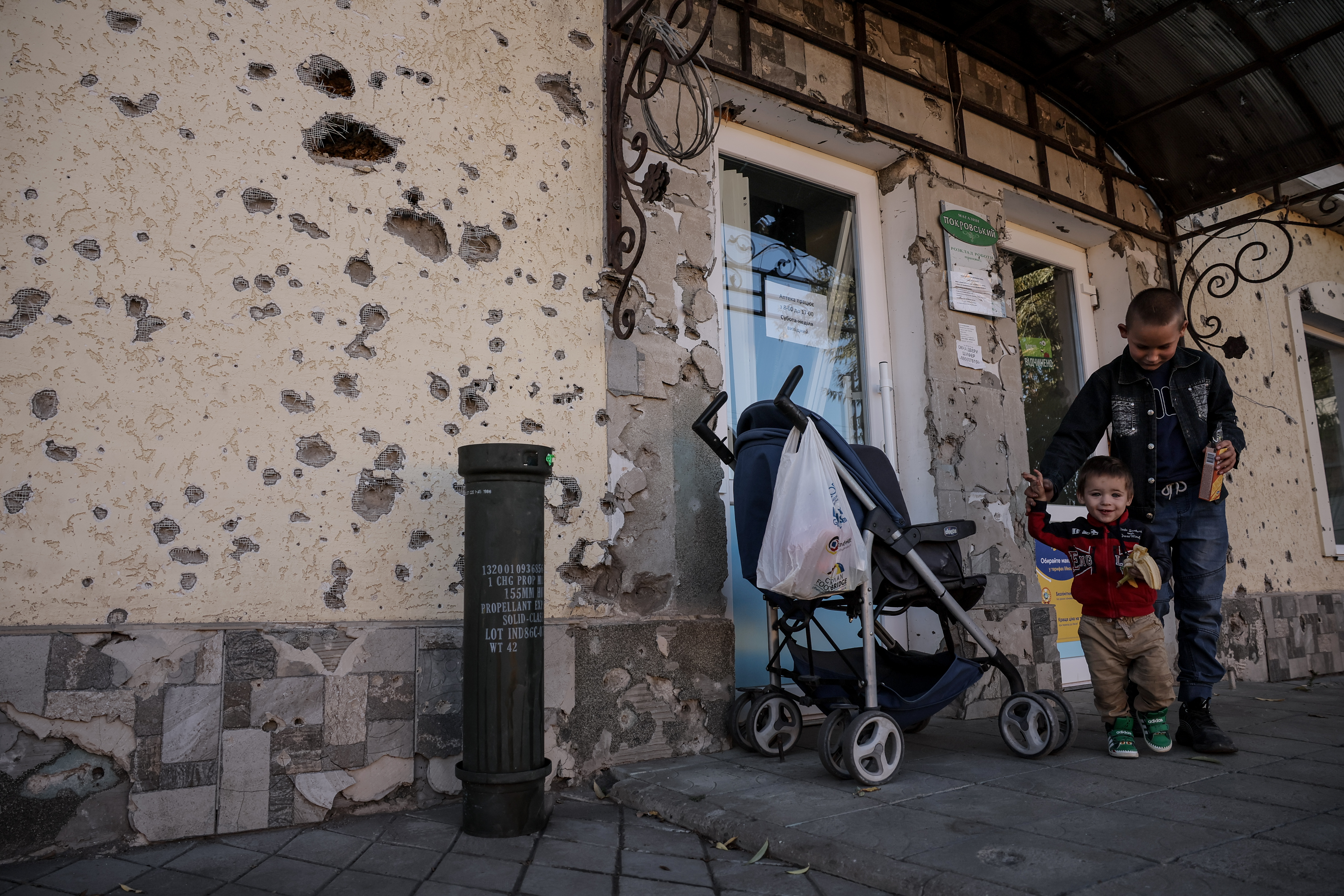 A boy and his baby cousin outside a shop in the southern Kherson region. The wall is pockmarked with bullet holes.