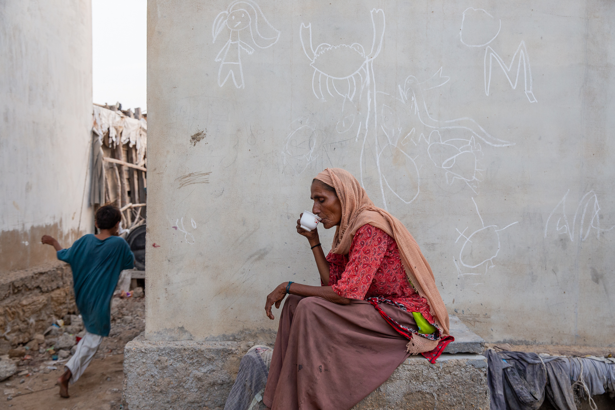 A photo of a woman sitting down and drinking something from a mug with a child running on the left.