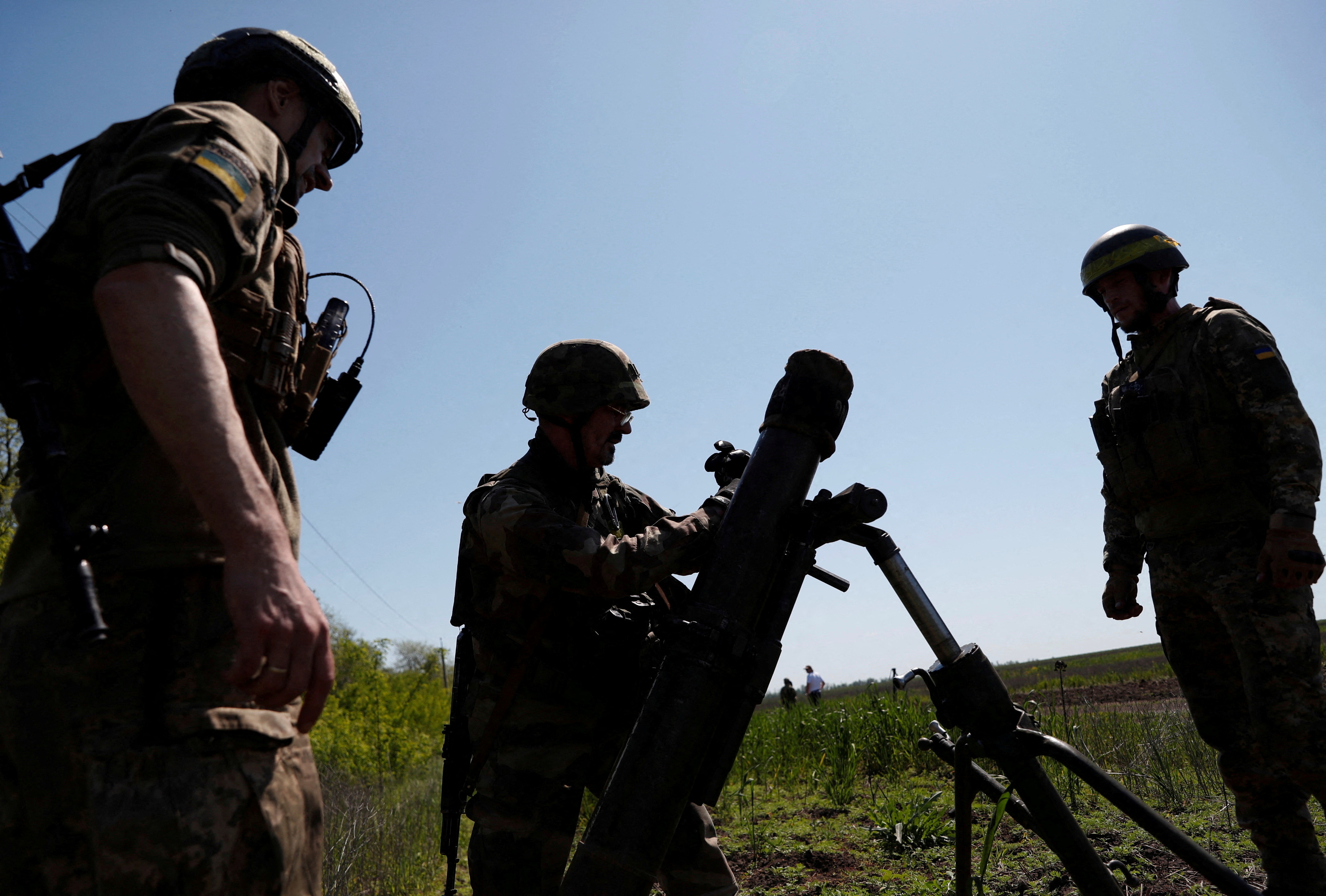 Three members of Ukraine's 128th Mountain Assault Brigade setting up an 120mm mortar during training in May. They are silhouetted against a blue sky. The mortar is in the middle.