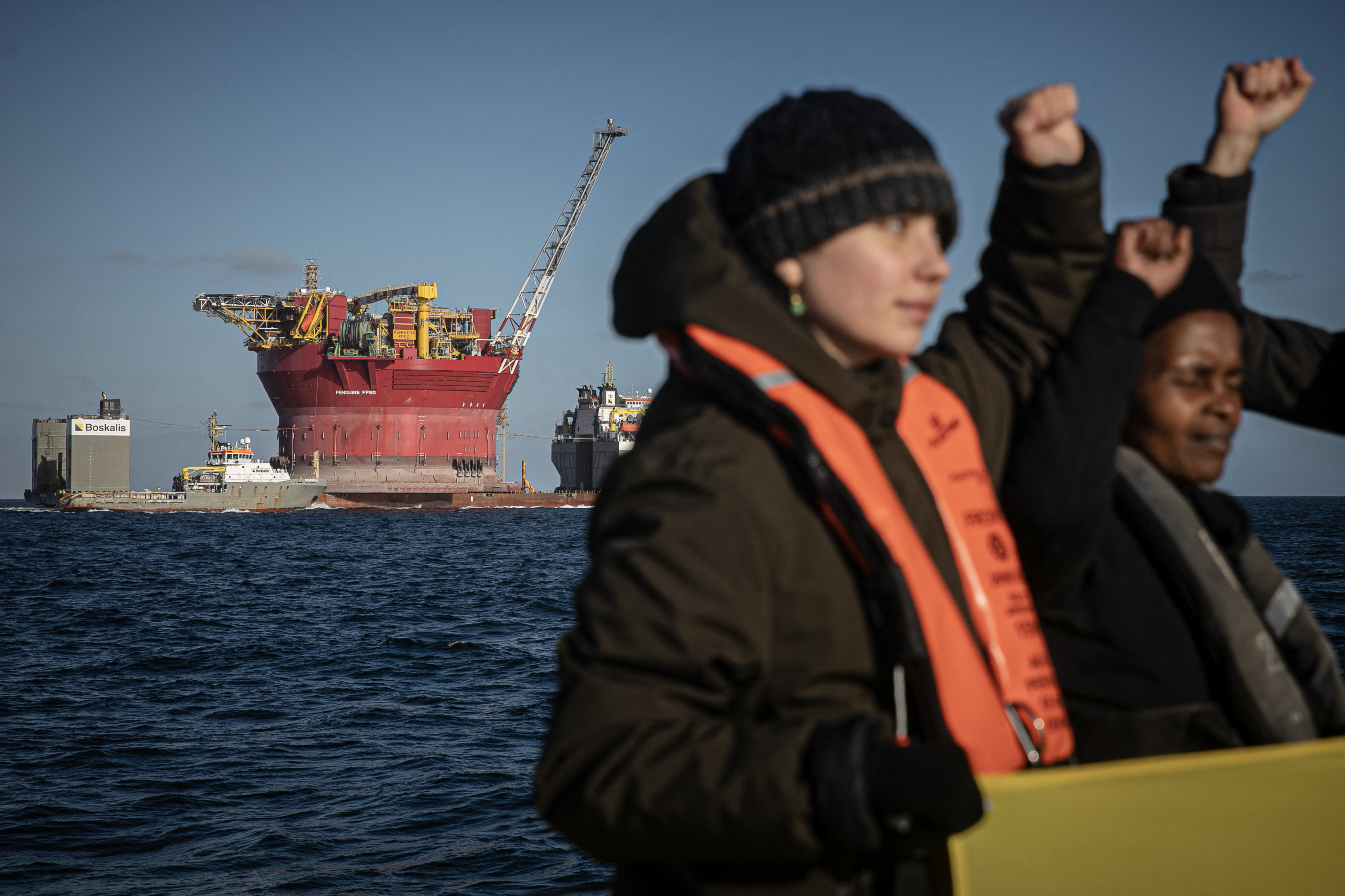 Protesters against oil punch the air. Behind them a platform is being towed by sea to expand an oil field.