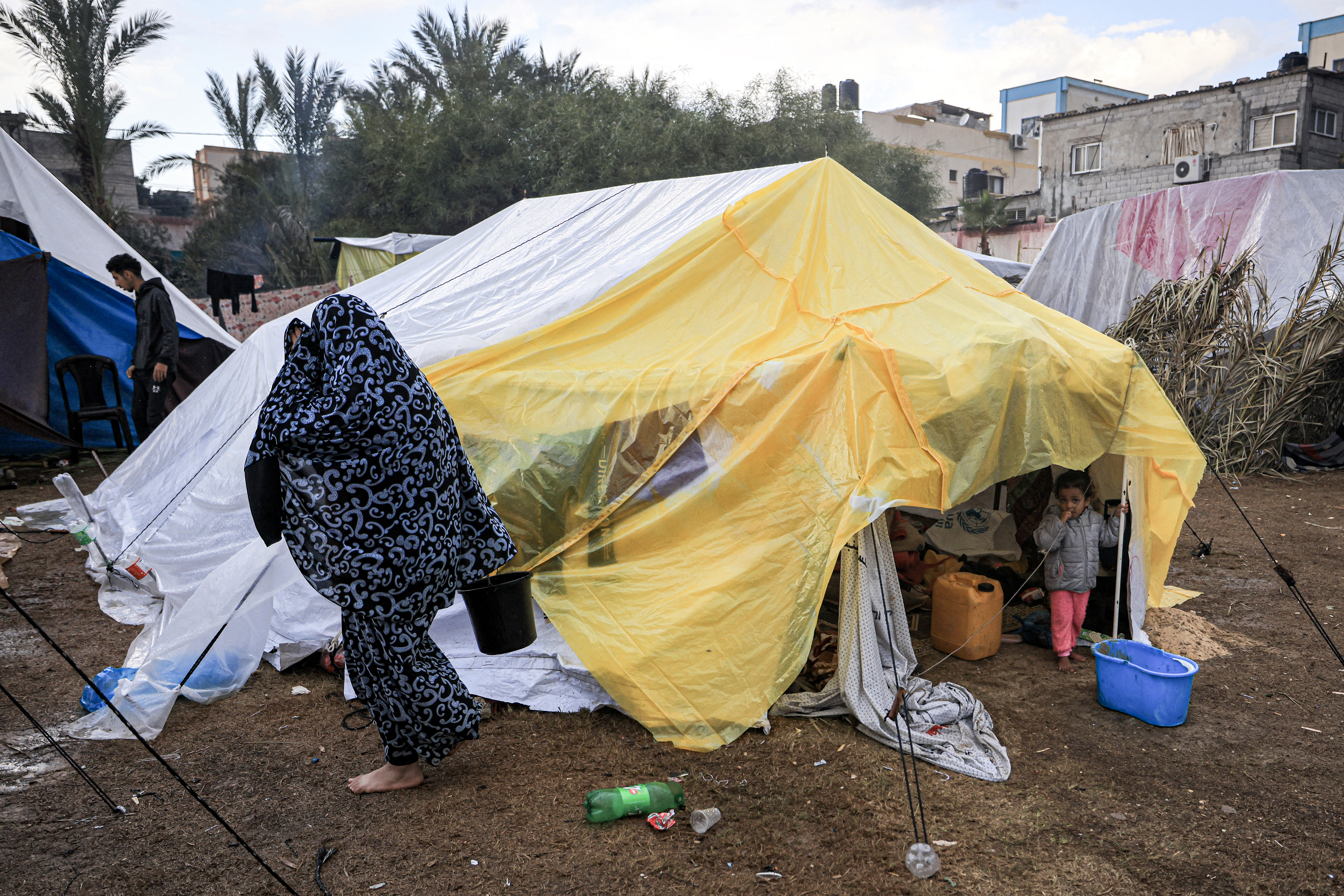 A woman walks past a girl stands outside the entrance of a tent covered with plastic insulation, pitched with others by Palestinians taking shelter from Israeli bombardment around Nasser Hospital, in Khan Yunis