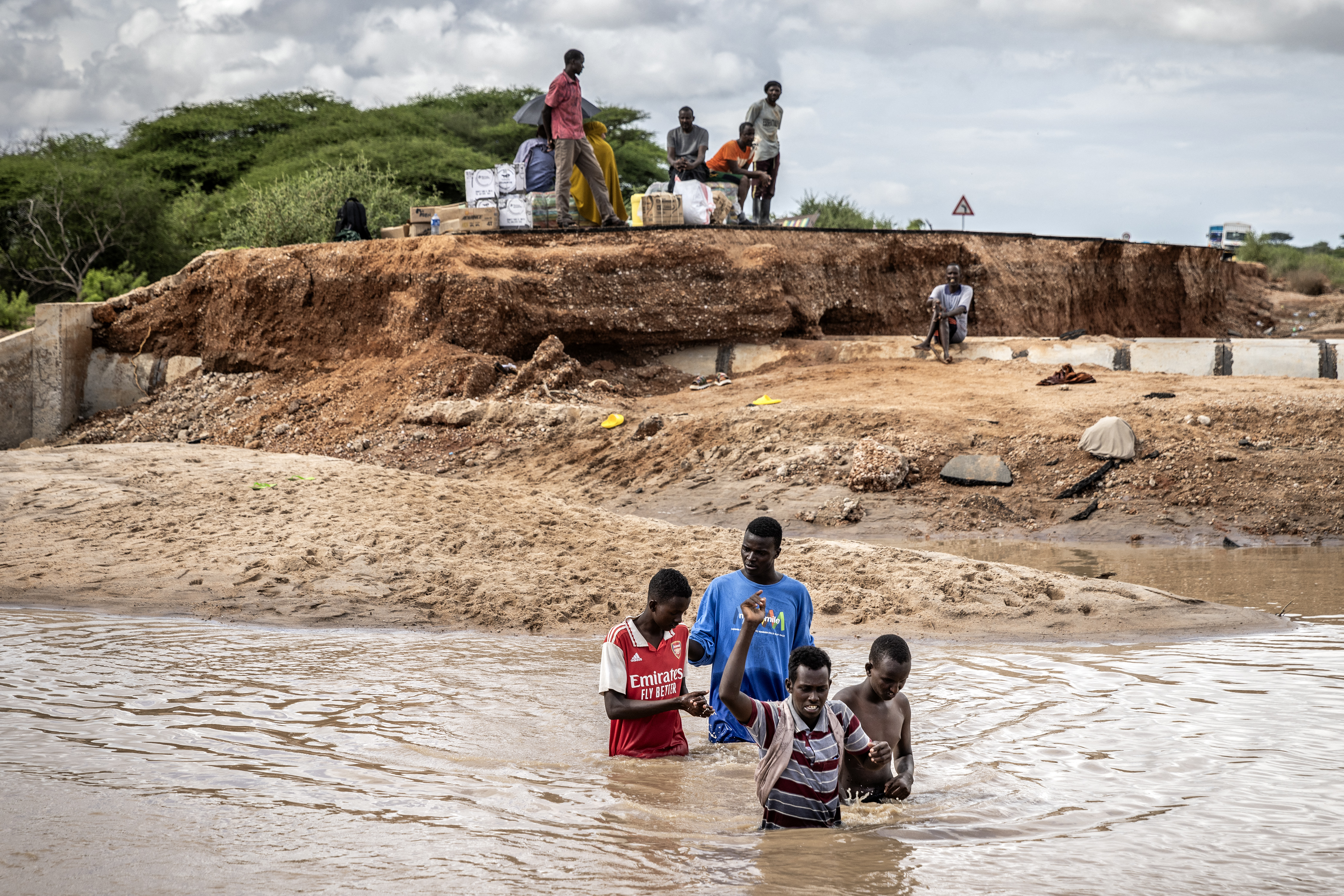 People gather waiting for goods on top of the section of a road destroyed by flash floods as other try to cross the waters at the Garissa-Wajir Road in Maalimin.