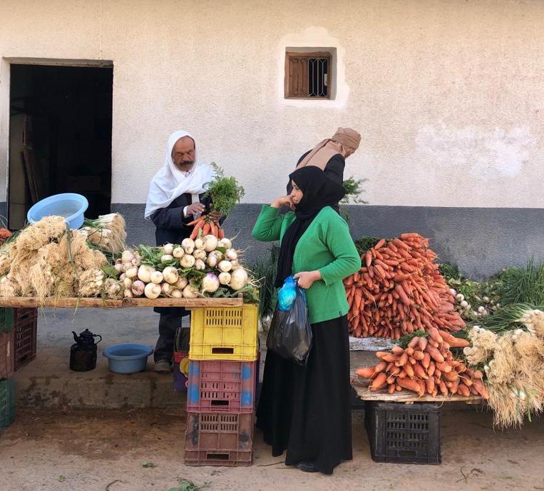 A photo of someone at an outdoor market buying vegetables with two manning the stall with piles of vegetables.