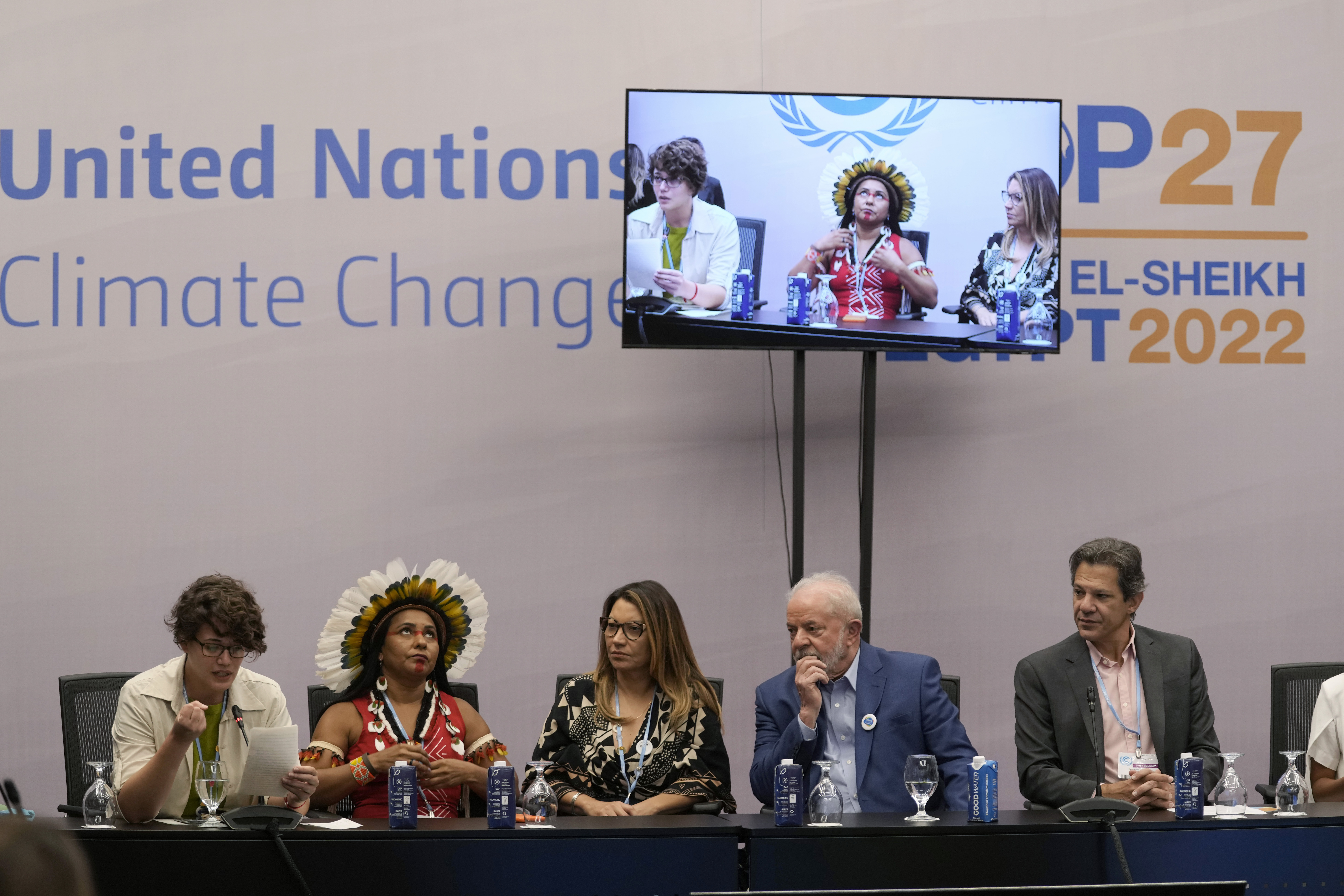 Indigenous leader Puyr Tembe, wearing a crown of radiating feathers, sits on a United Nations Climate Change panel with Brazilian President Lula da Silva, who is dressed in a blue suit. A TV screen broadcasts their remarks above them. 