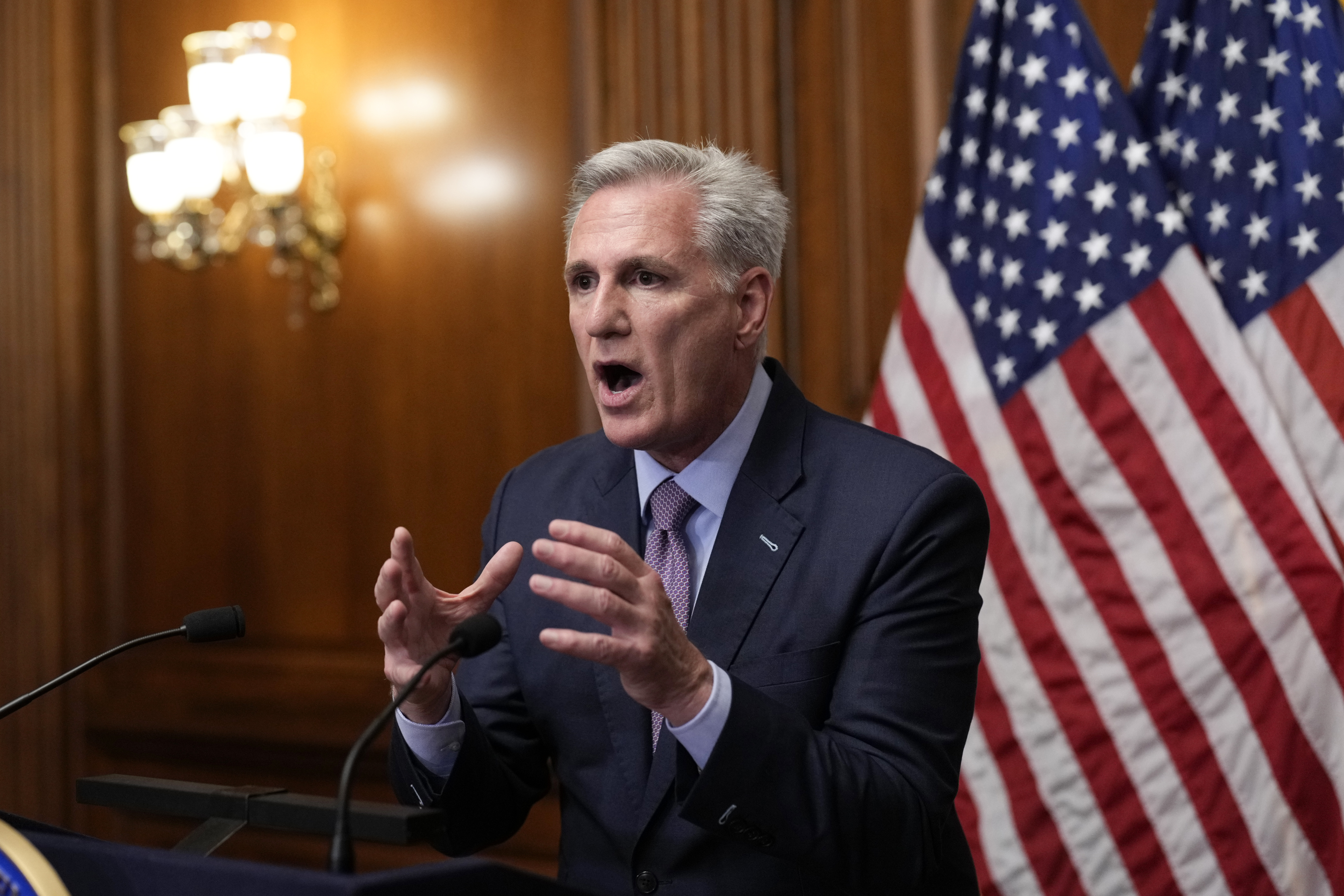 Kevin McCarthy gestures with both hands outstretched in a wood-paneled room decorated with US flags.