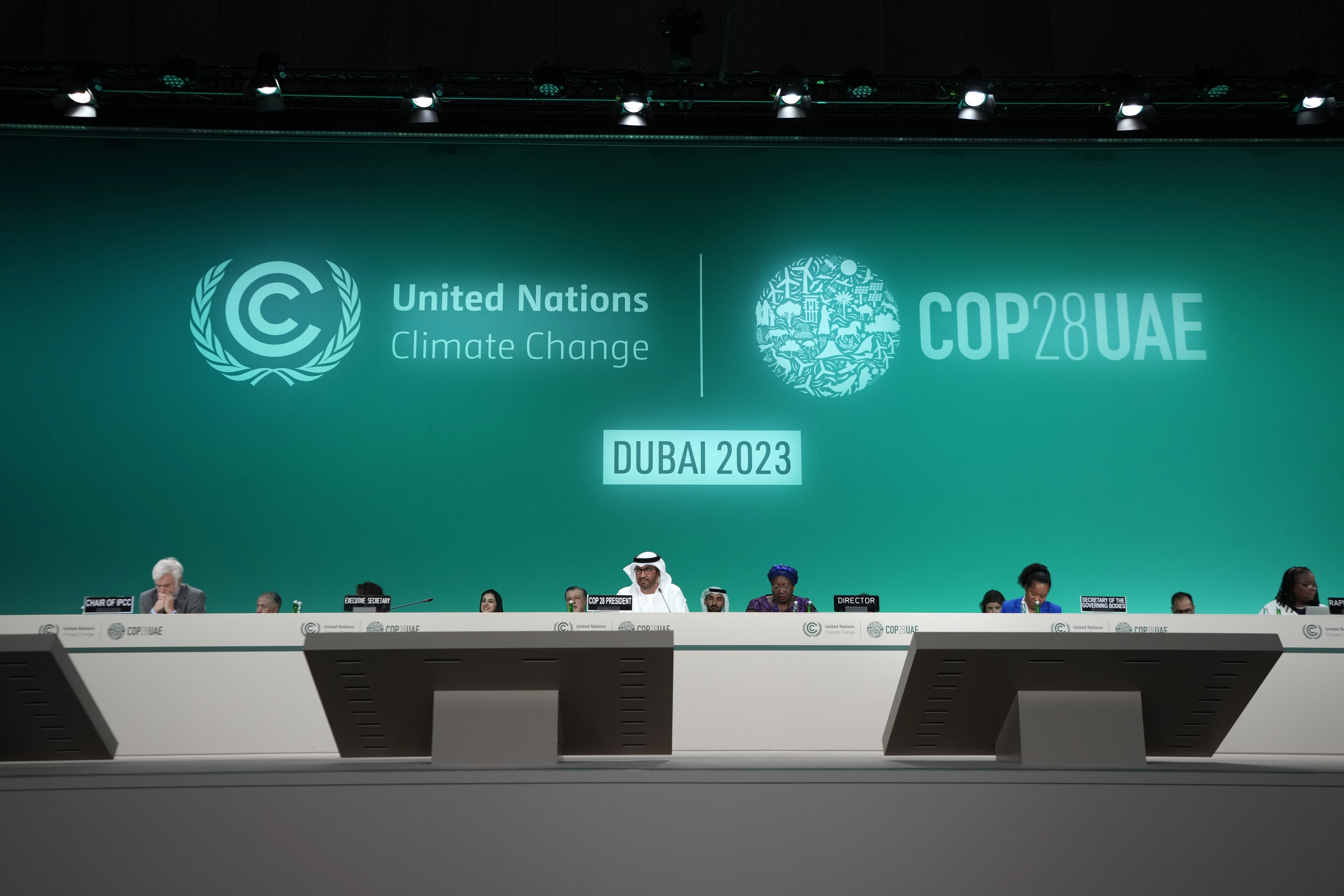 A panel table sits along the length of a stage in Dubai, behind which officials sit. The background of the stage is a green screen with logos and slogans like "Dubai 2023," "United Nations Climate Change" and "COP28 UAE."
