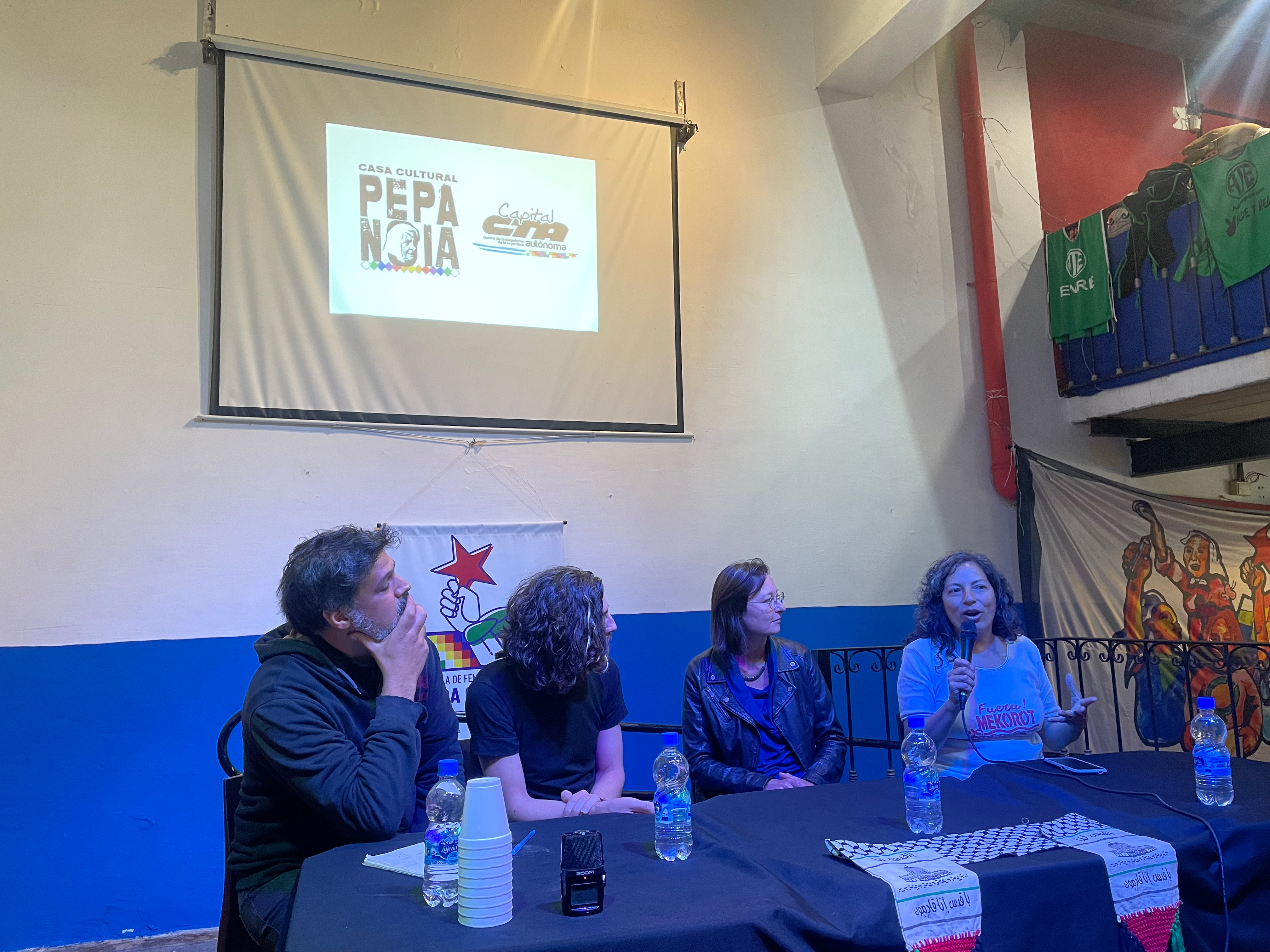 A panel of speakers sits at a table, where Silvia Ferreyra speaks into a microphone. Behind them, a projection on a screen mounted on the wall indicates they are in the Casa Cultural Pepa Noia.