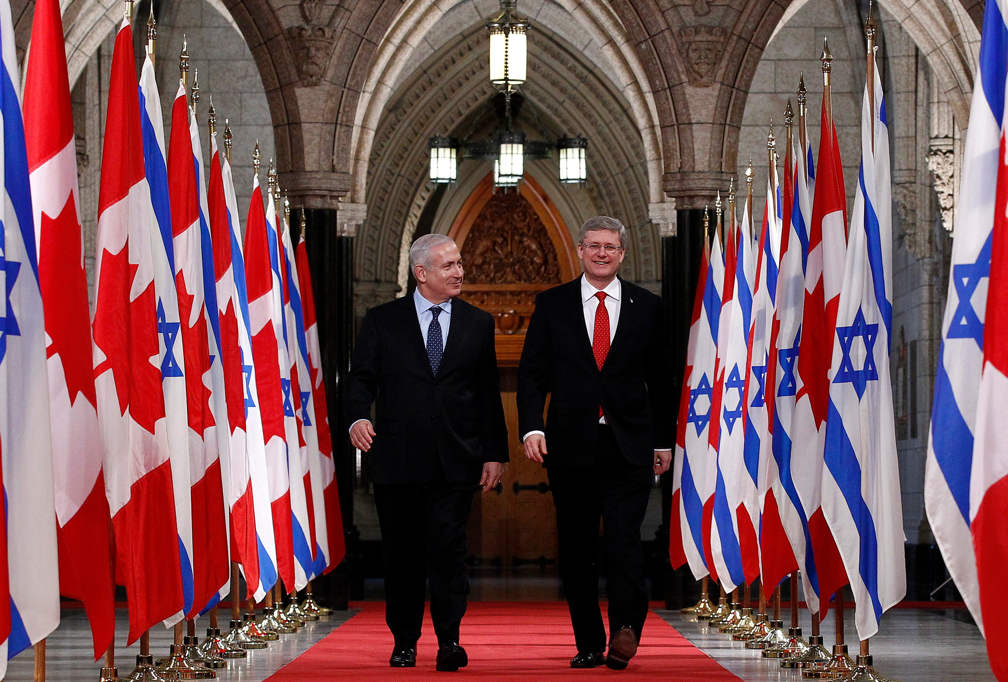 Stephen Harper walks side by side with Benjamin Netanyahu, as they stroll down a red carpet surrounded on either side by Canadian and Israeli flags.