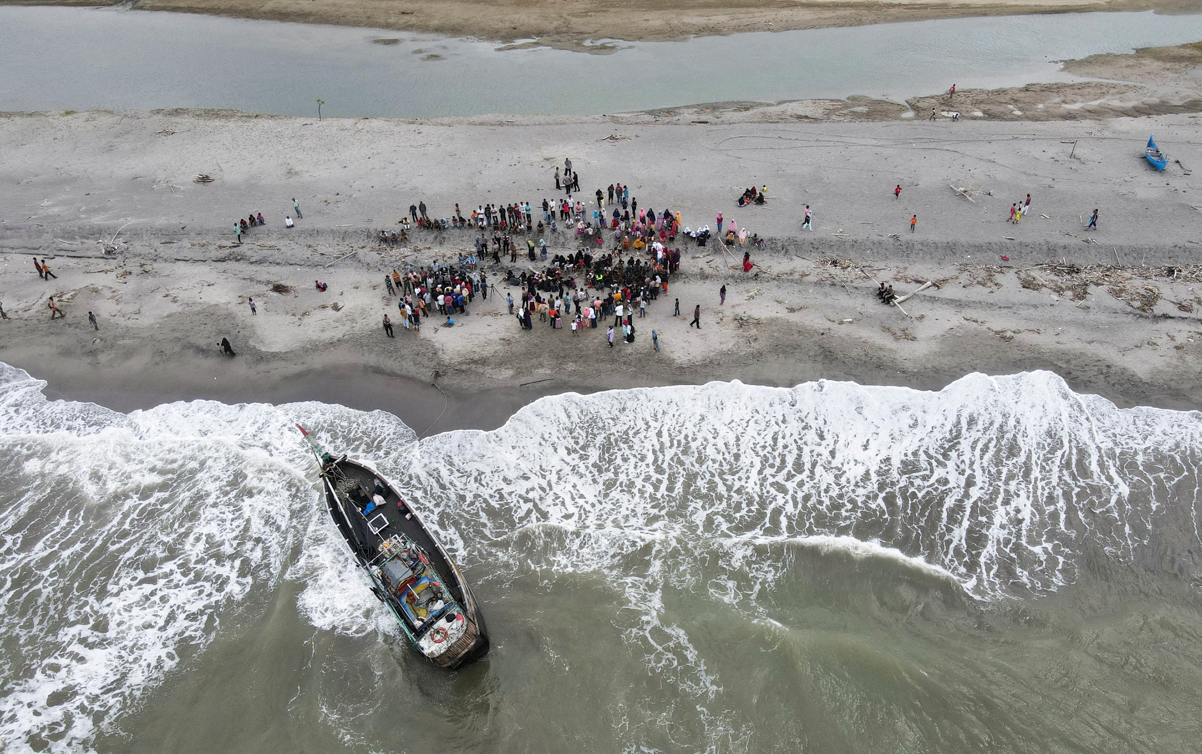 An aerial view of a boat amid waves crashing on the beach and dozens of passengers ashore.