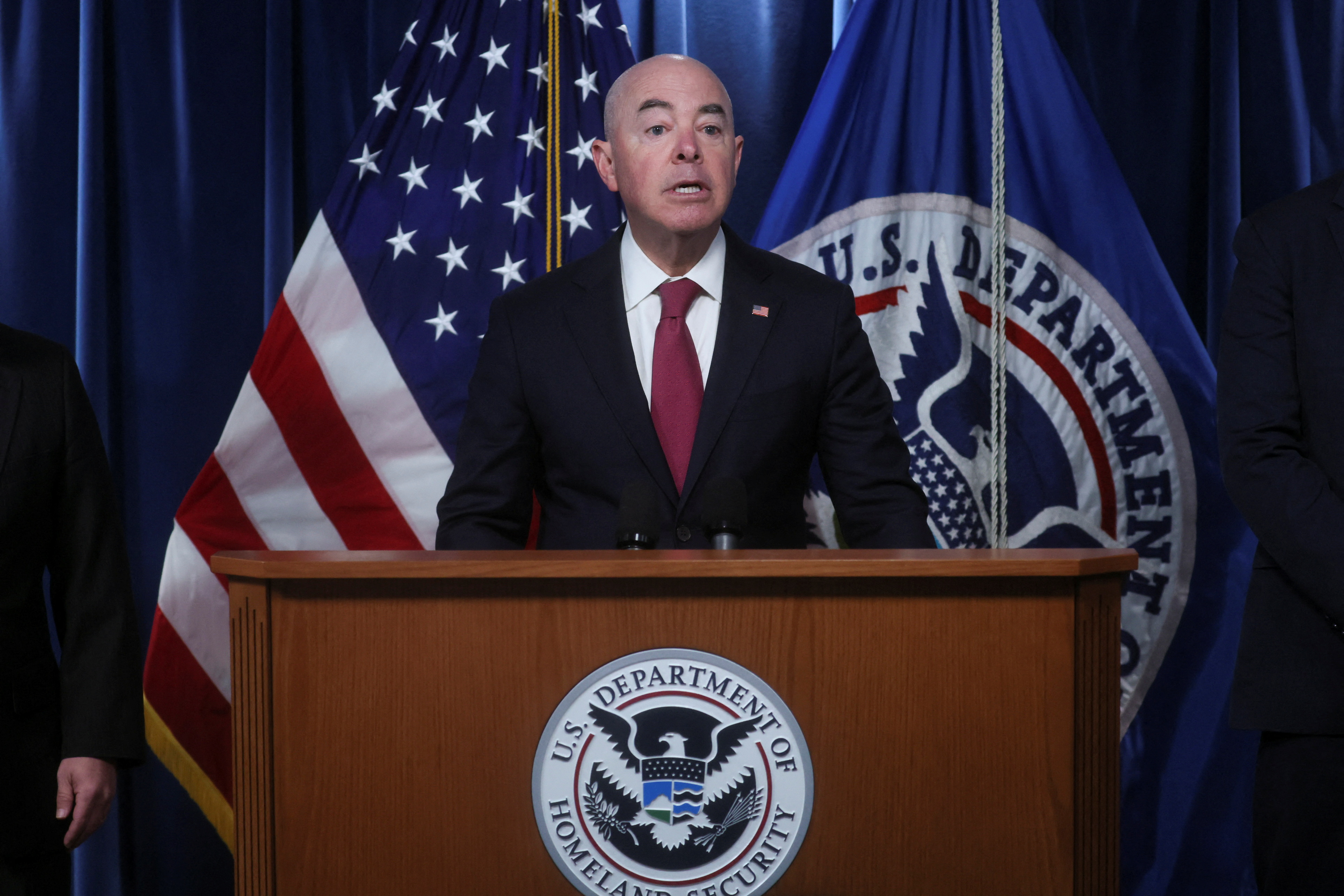 Alejandro Mayorkas stands in behind a podium, with a US flag and a flag for the Department of Homeland Security behind him.