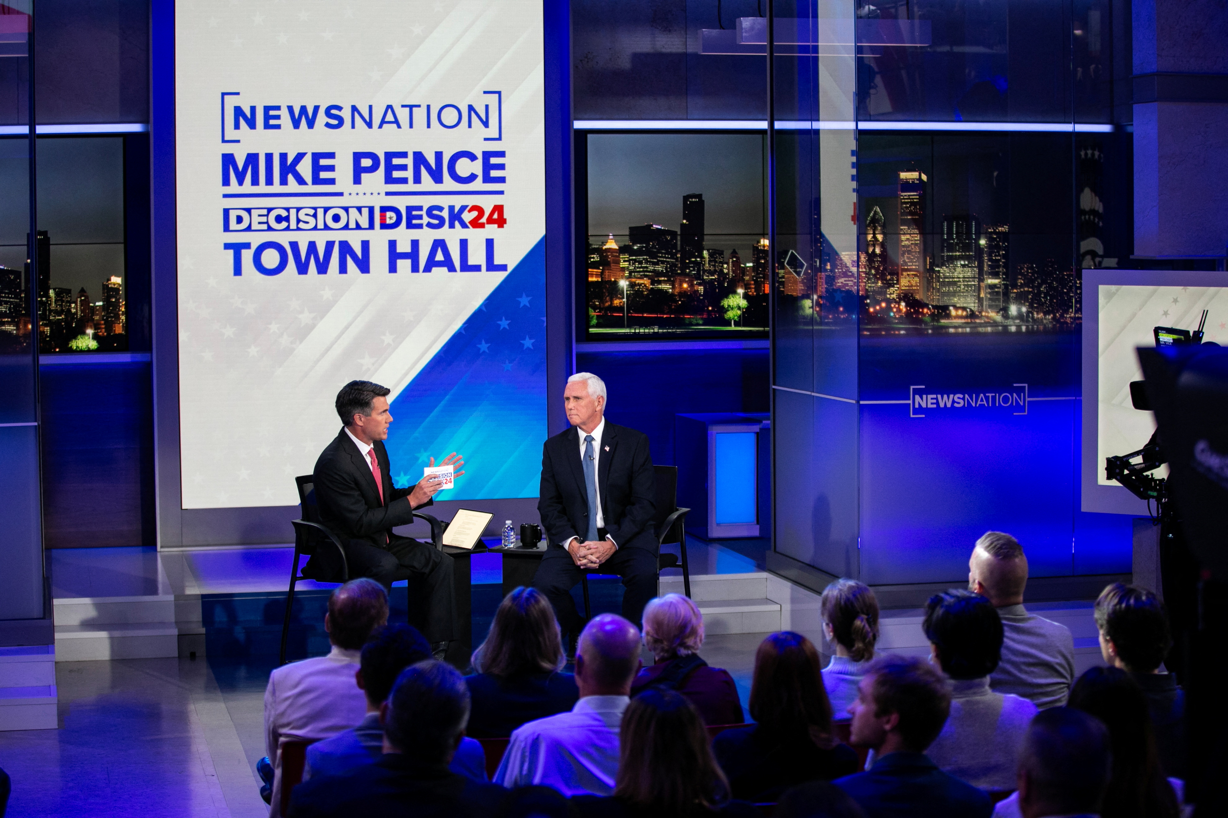 Mike Pence sits opposite a host on a "town hall" stage in front of an audience. Behind them is a screen with the words, "NewsNation Mike Pence Election Desk town hall."