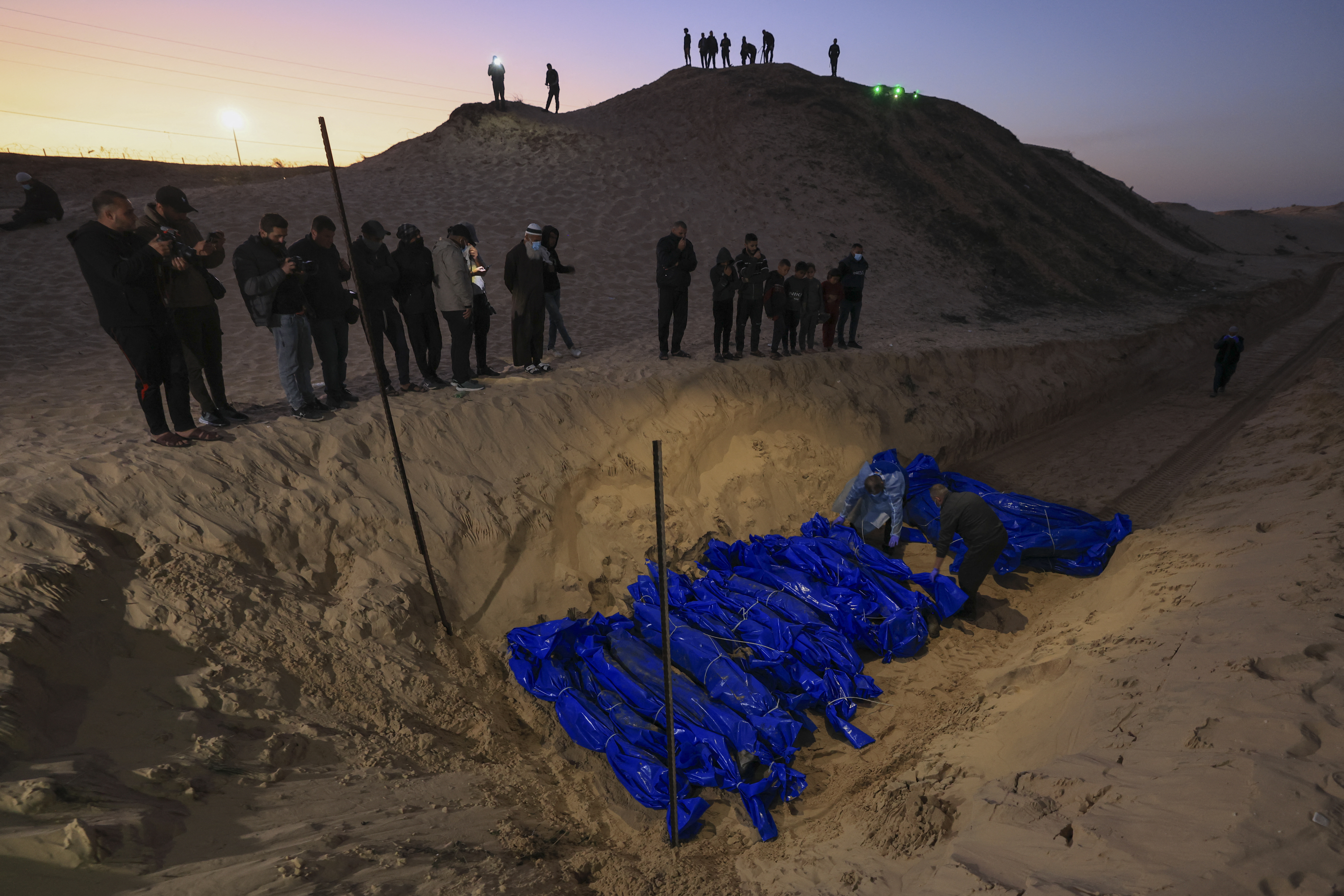 People look on as the shrouded bodies of Palestinians killed in nothern Gaza, that were taken and later released by Israel, are burried in a mass grave in Rafah, on the southern Gaza Strip on December 26, 2023, amid ongoing battles between Israel and the militant group Hamas. (Photo by Mahmud HAMS / AFP)