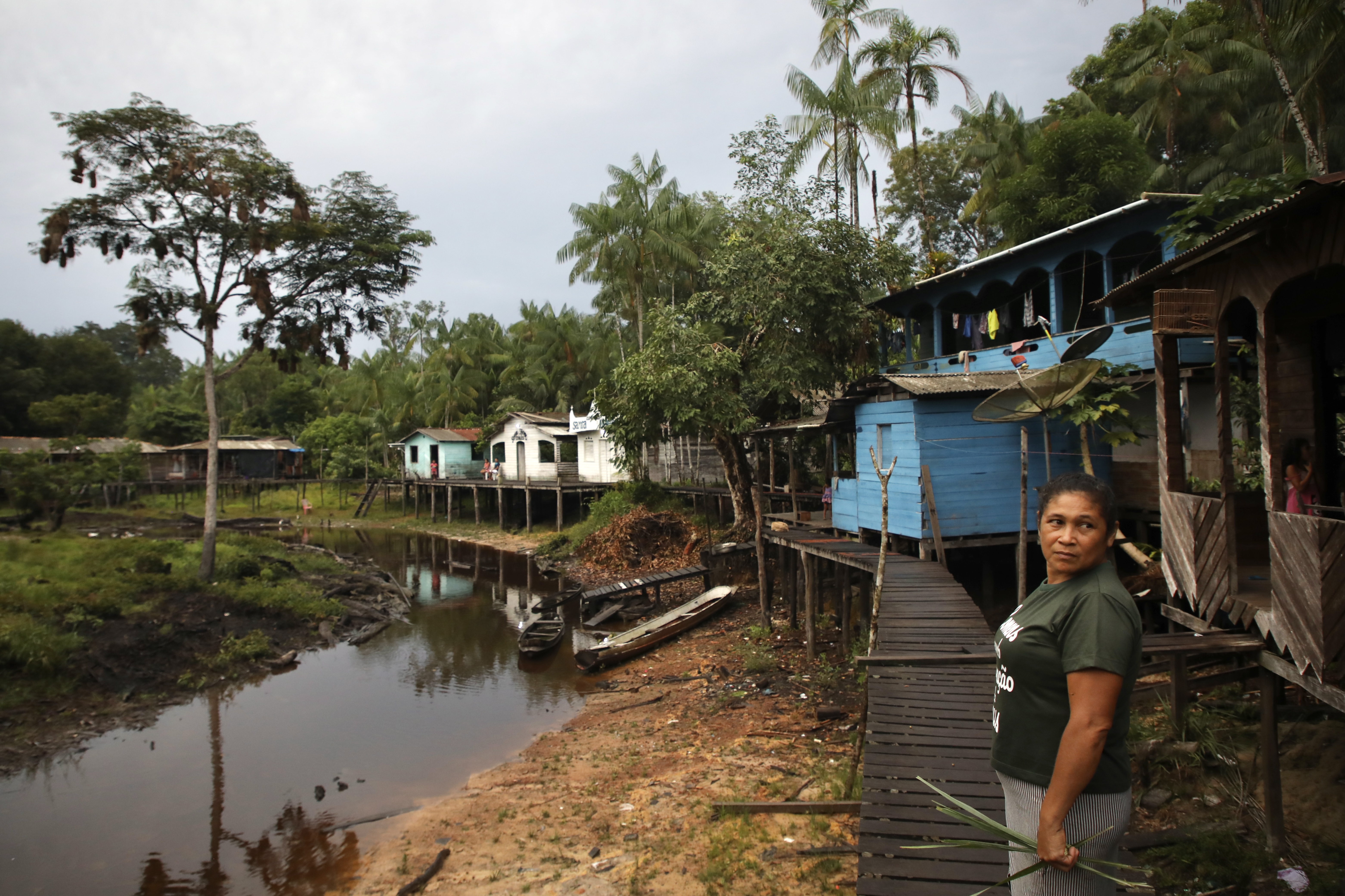 A woman stands on a walkway made of wooden planks that runs along a dried-up river. Houses on stilts sit along the river bank.