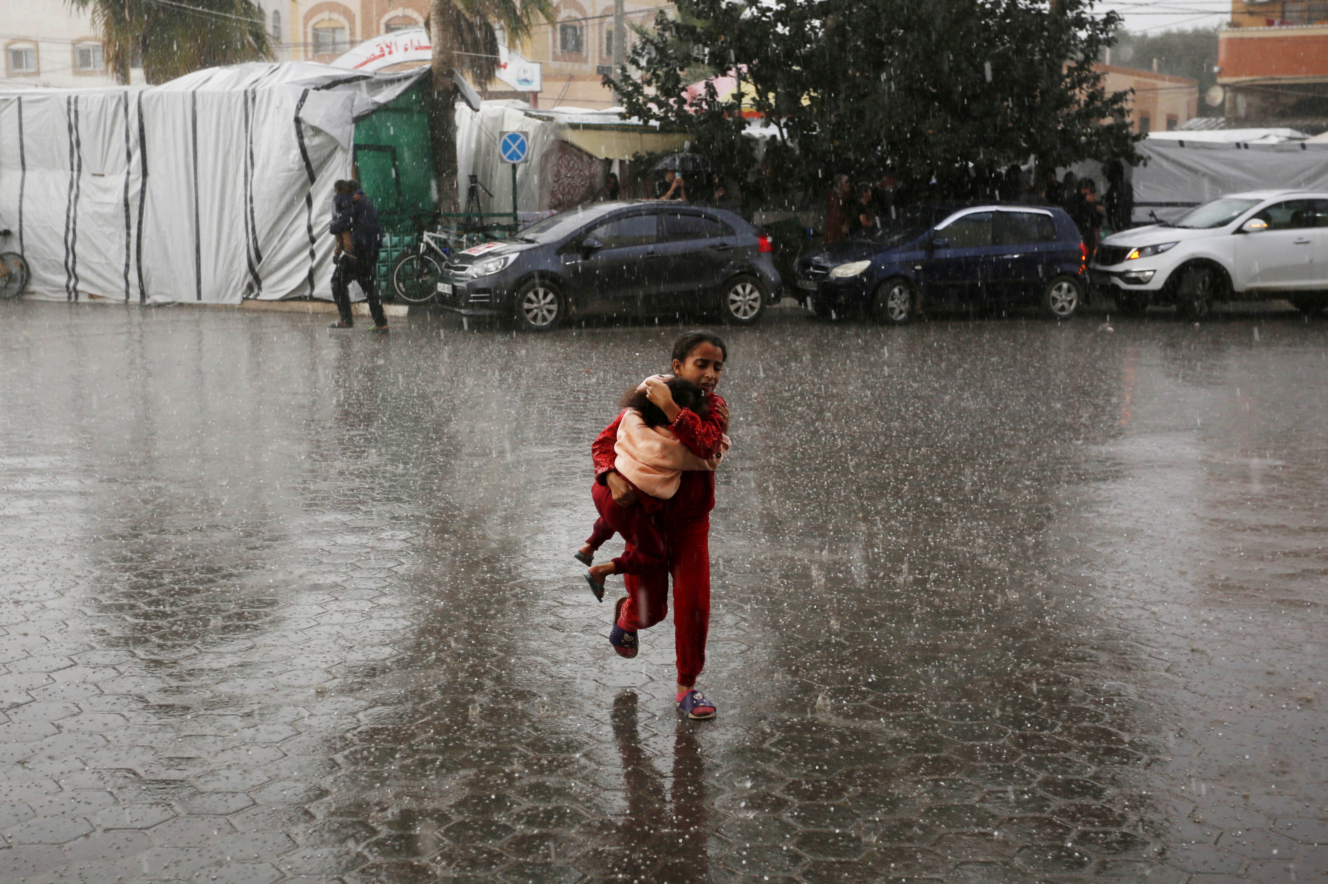 A Palestinian girl, trying to protect her sister from getting wet, walks under heavy rain as Palestinians who took shelter in the Aqsa Martyrs Hospital and its surroundings in Deir Al-Balah city of the Gaza are struggling with the strong winds, heavy rain and floods affecting the region amid Israeli attacks.