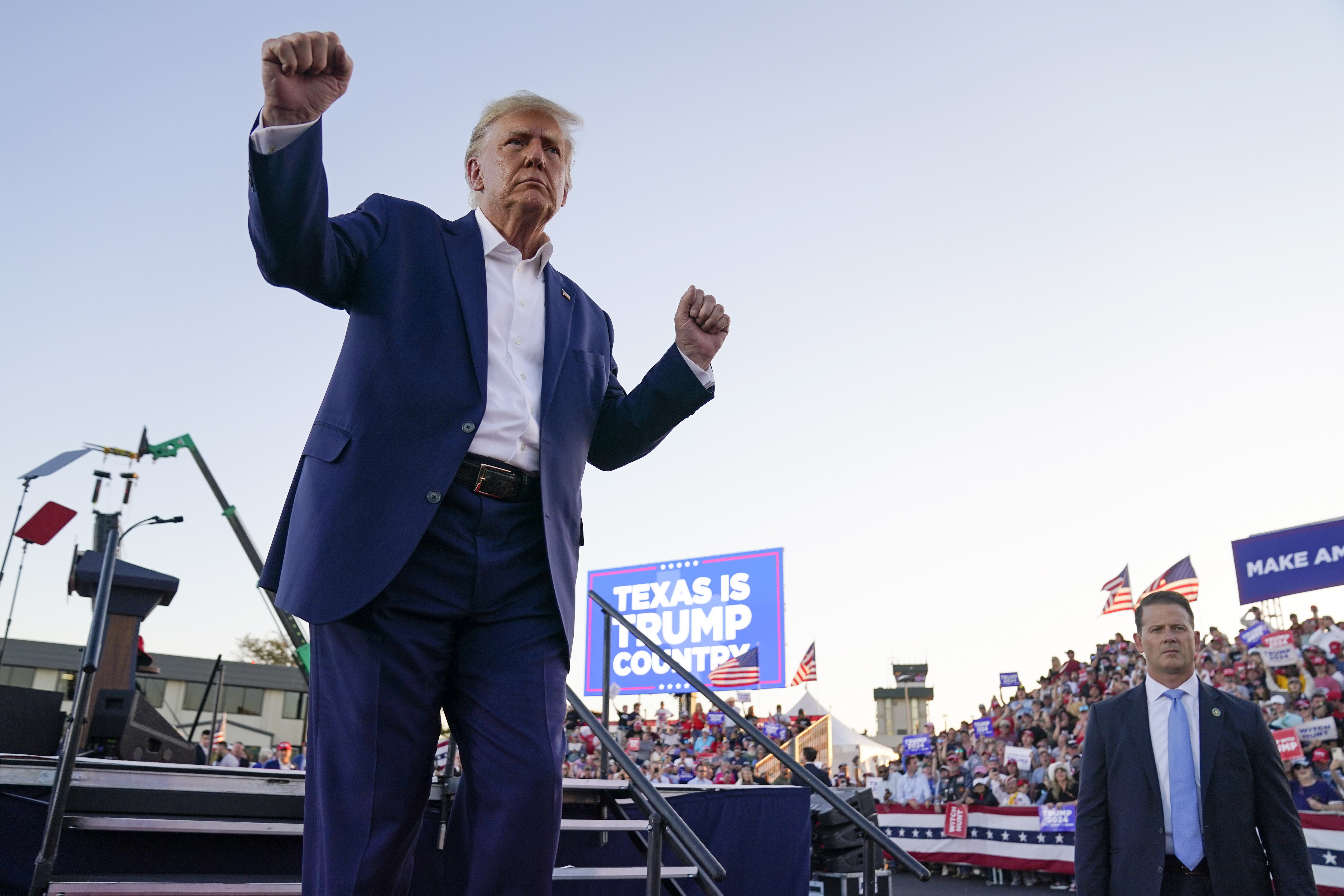 Trump lifts both his fists in the air at a campaign rally in Waco, Texas. Behind him, a screen reads, "Texas is Trump country," and crowds are visible.