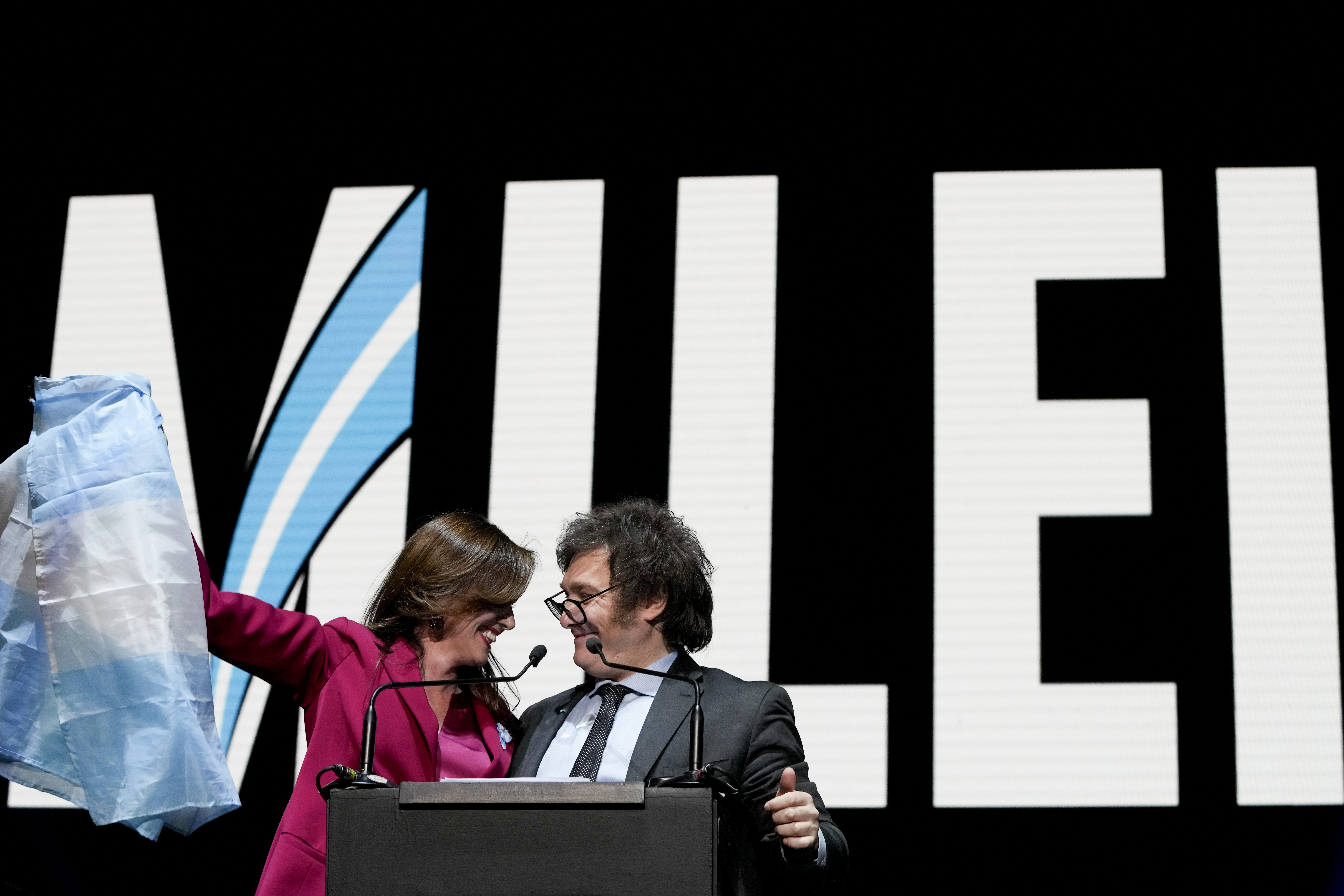 Javier Milei in a dark suit and Victoria Villarruel in a red suit stand in front of a screen with giant, capitalized letters that read, "Milei." Villarruel holds up an Argentine flag.
