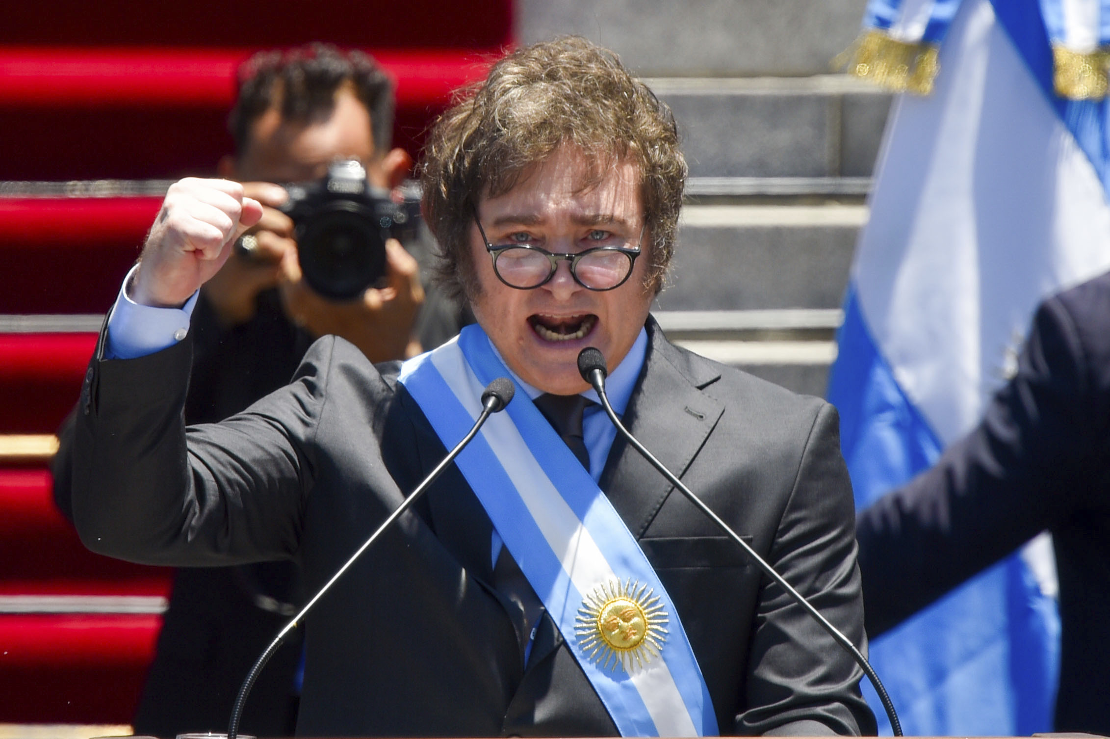 Javier Milei, shortly after being sworn in, speaks into the microphones at a podium outside Congress. He raises a fist as he speaks, wearing a dark suit, glasses and a sash in the colours of the Argentine flag. Behind him, a red carpet is visible on a set of granite steps.