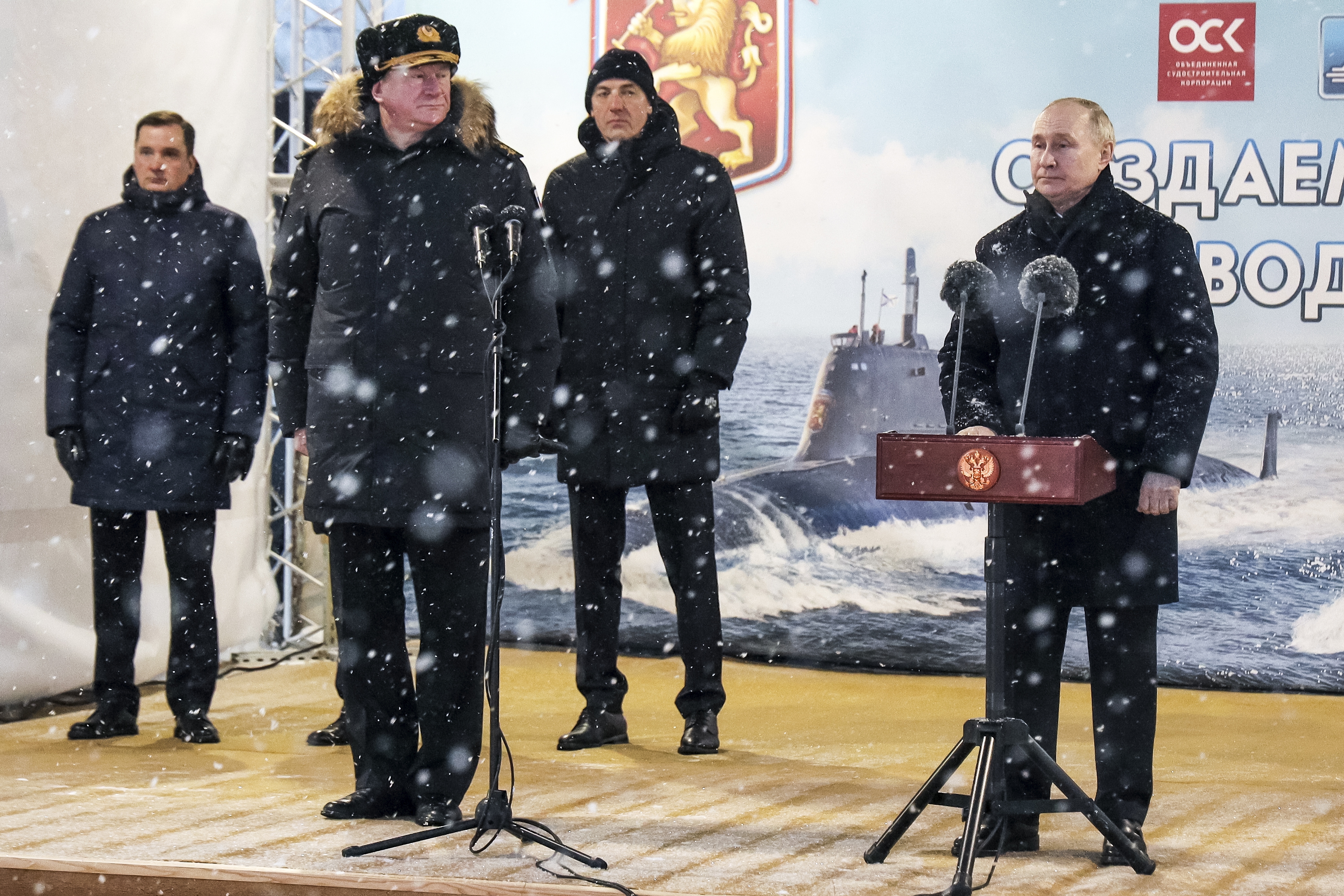 Putin speaking at an outdoor stage. Admiral Nikolai Yevmenov, Commander-in-Chief of the Russian Navy, is to his right. Other senior naval officers are also on stage. It is snowing.