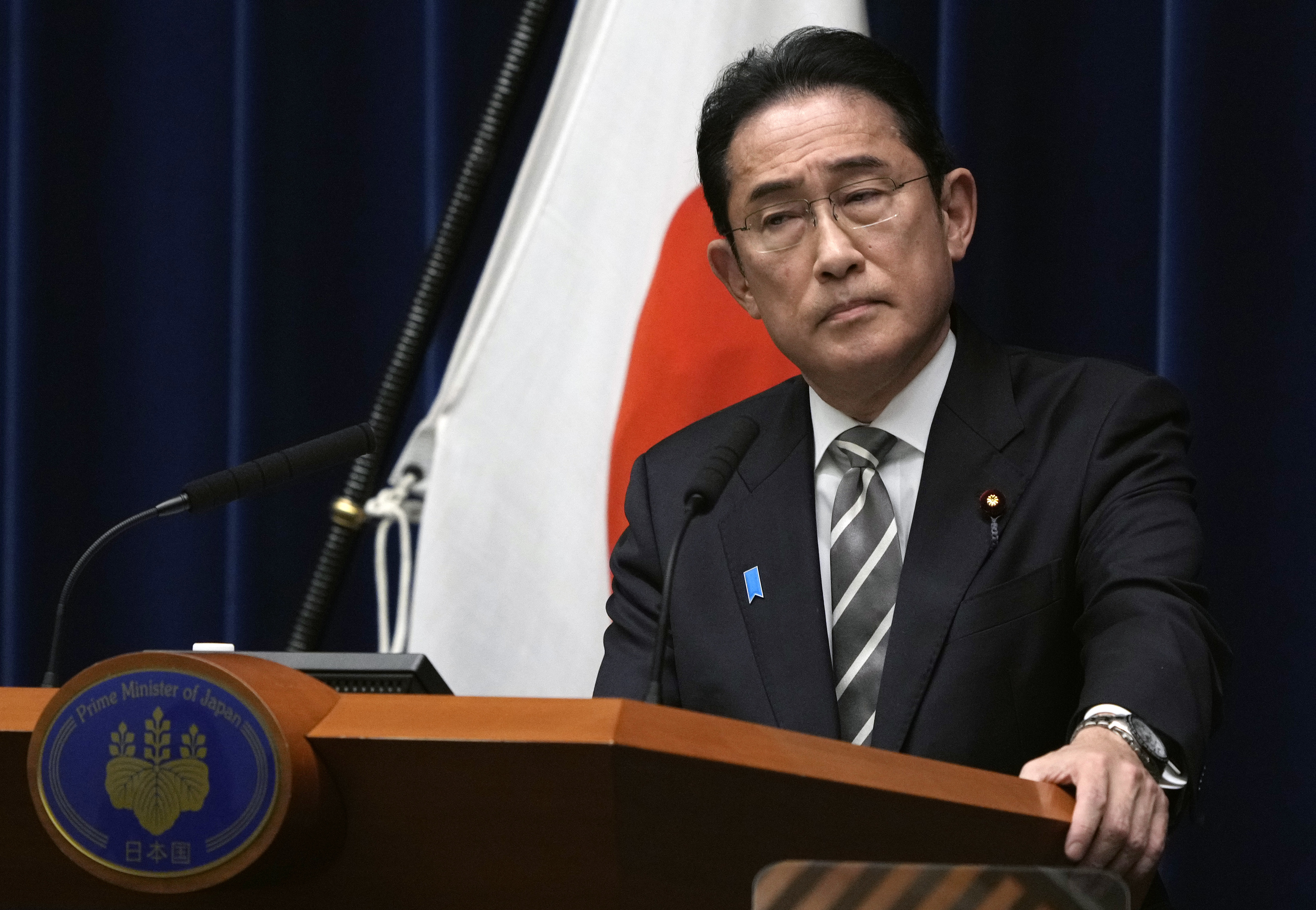 Japanese Prime Minister Fumio Kishida. He is standing at a lectern in front of a Japanese flag. He looks serious.