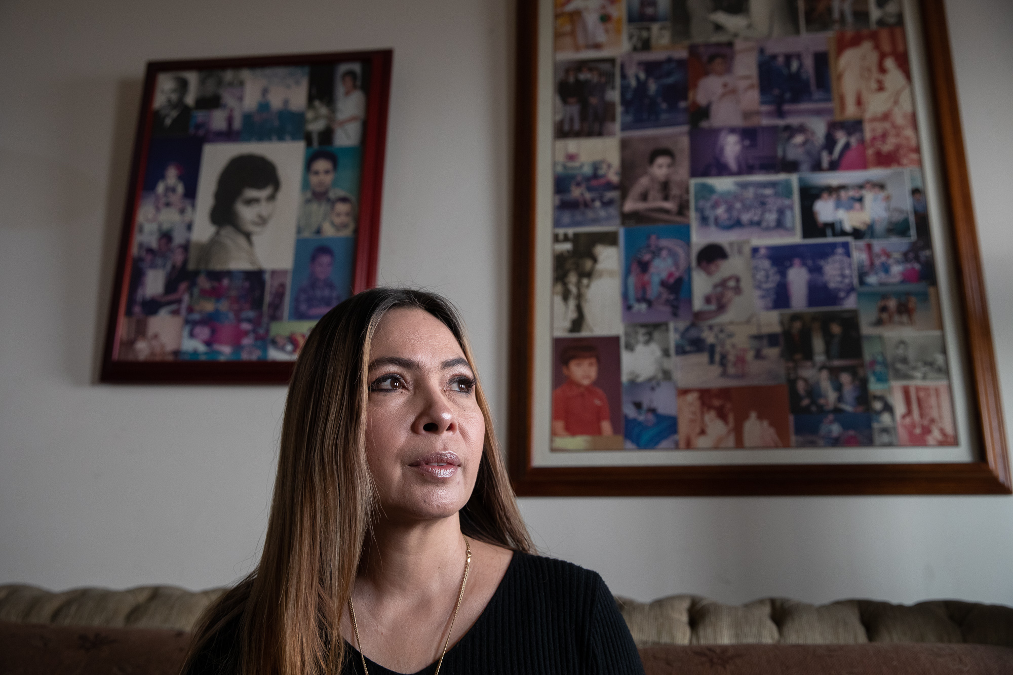 Decnis Daiz sits on a couch. Behind her are framed collages of family photos.