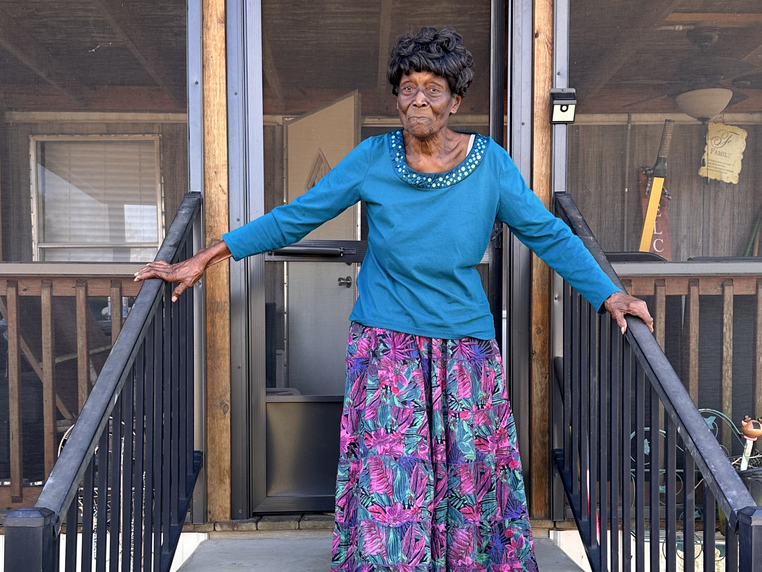 An elderly woman in a blue top and colourful skirt stands outside a building