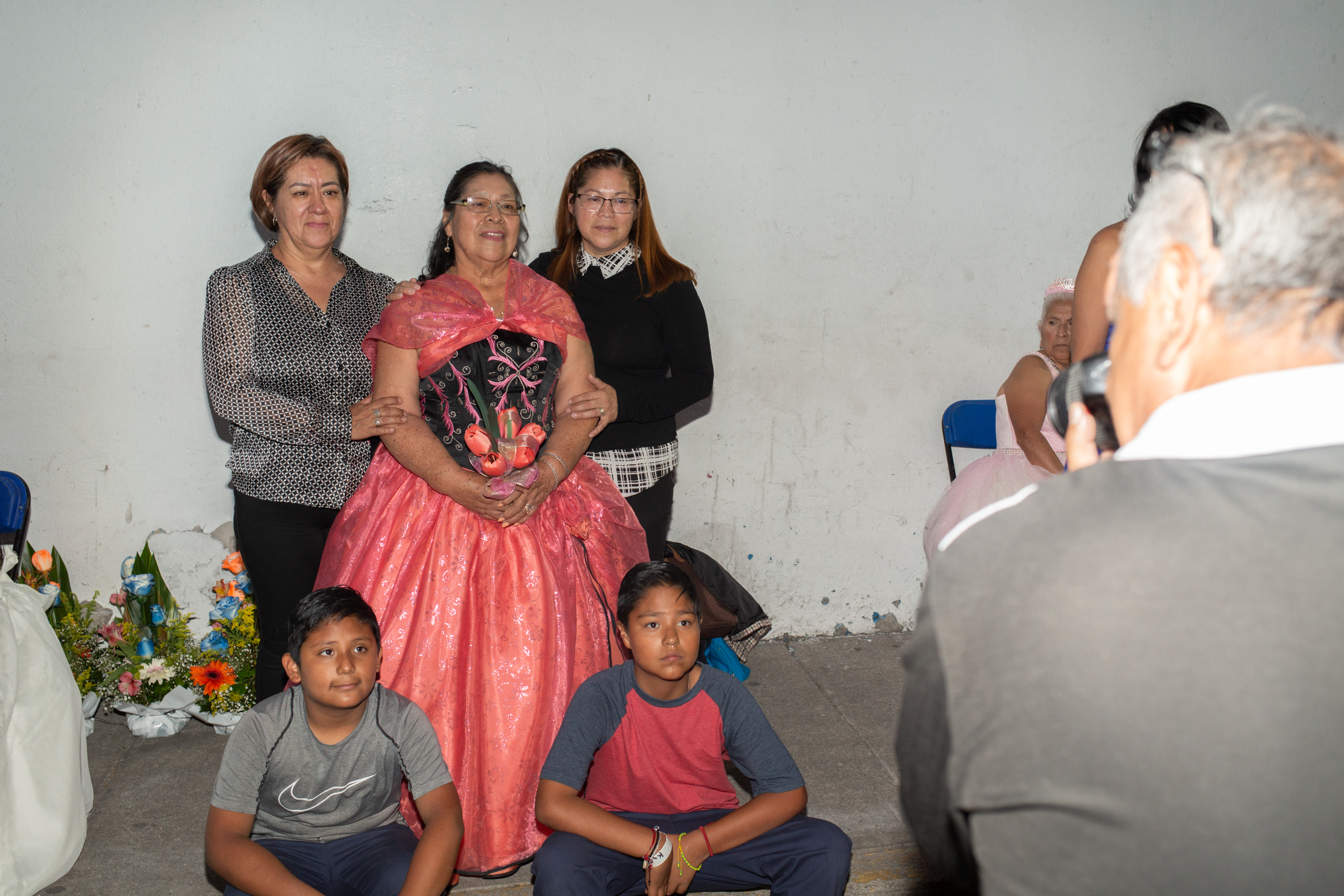 A woman in a hot pink ballgown with a blue bodice carries a bouquet of matching pink tulips as she is surrounded by her family, two women on either side of her and a pair of young boys at her feet. Together, they pose for a photo.