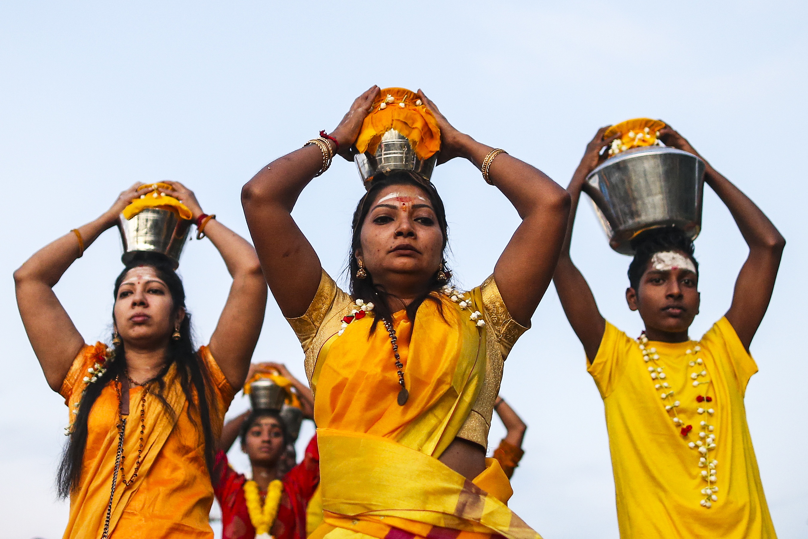 Two women and a young man carrying pots of milk on their heads. They are dressed in orangey/yellow clothing.