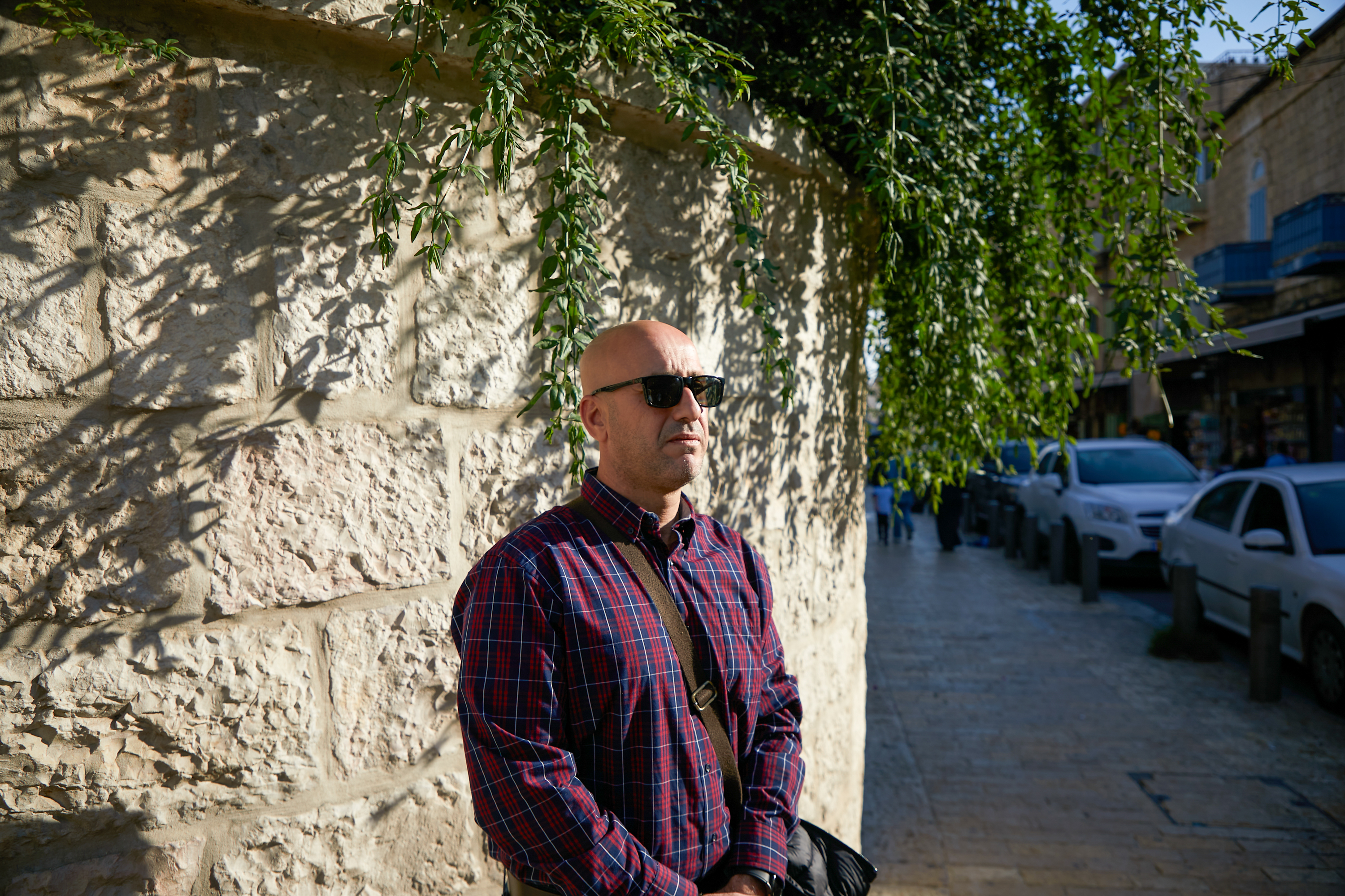 Shadi in a plaid shirt and sunglasses standing in the sun near a stone wall