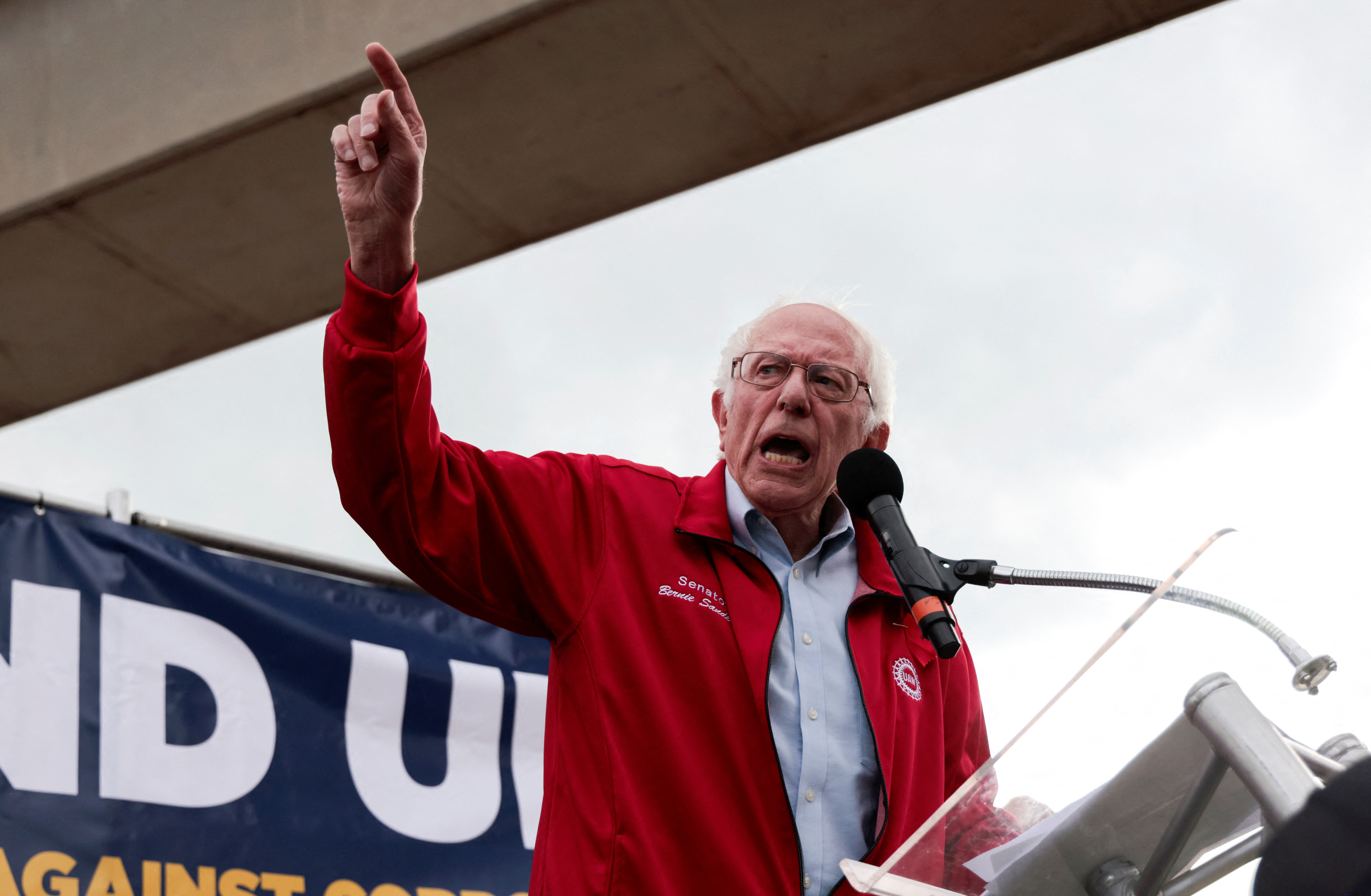 Bernie Sanders, wearing a blue collared shirt and a red jacket, points upward with a finger as he speaks into a microphone outside at a rally.