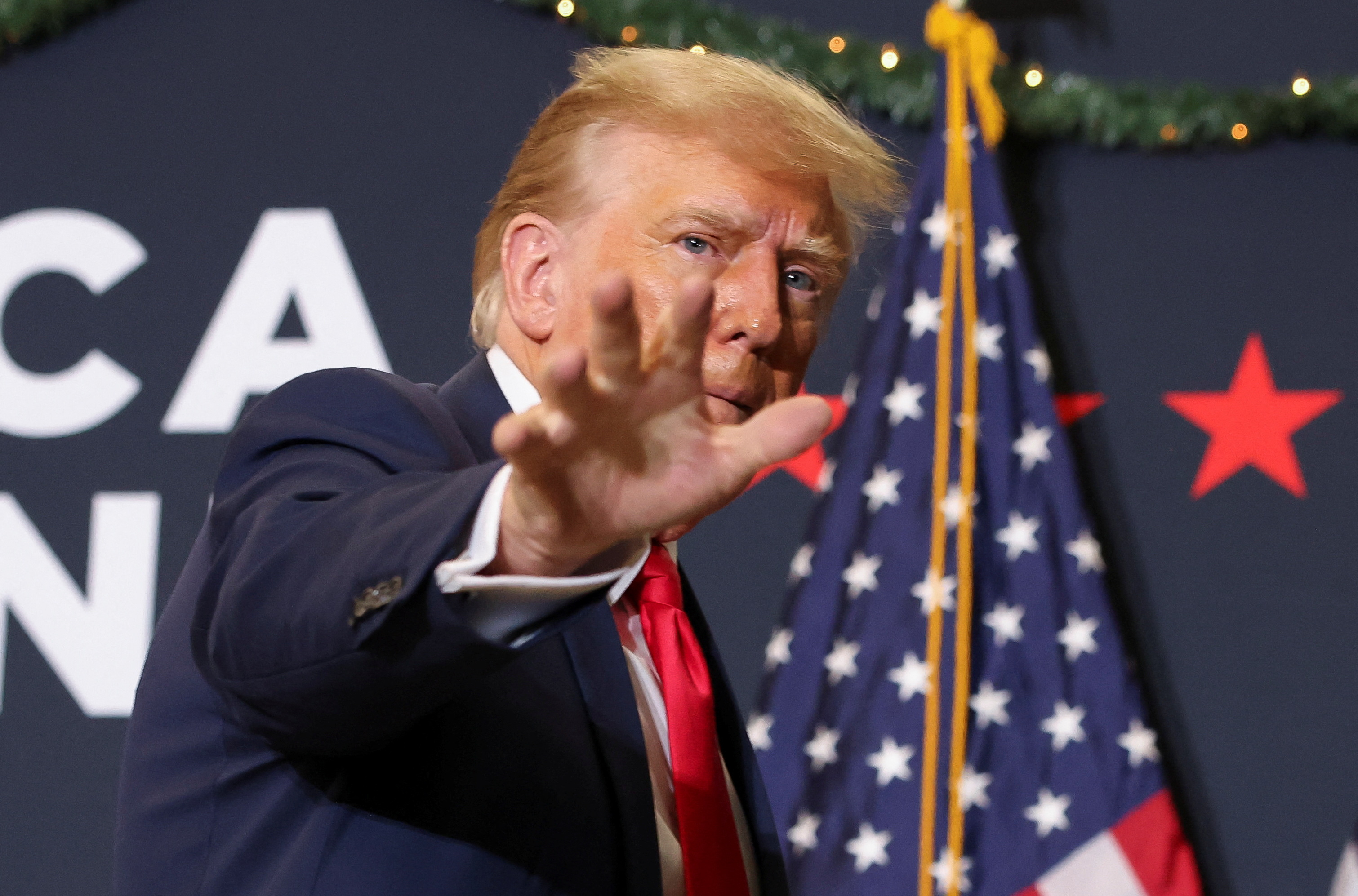 Donald Trump extends his hand, his fingers outstretched, toward the crowd at a December 2023 campaign rally. Behind him is a US flag and a campaign banner.