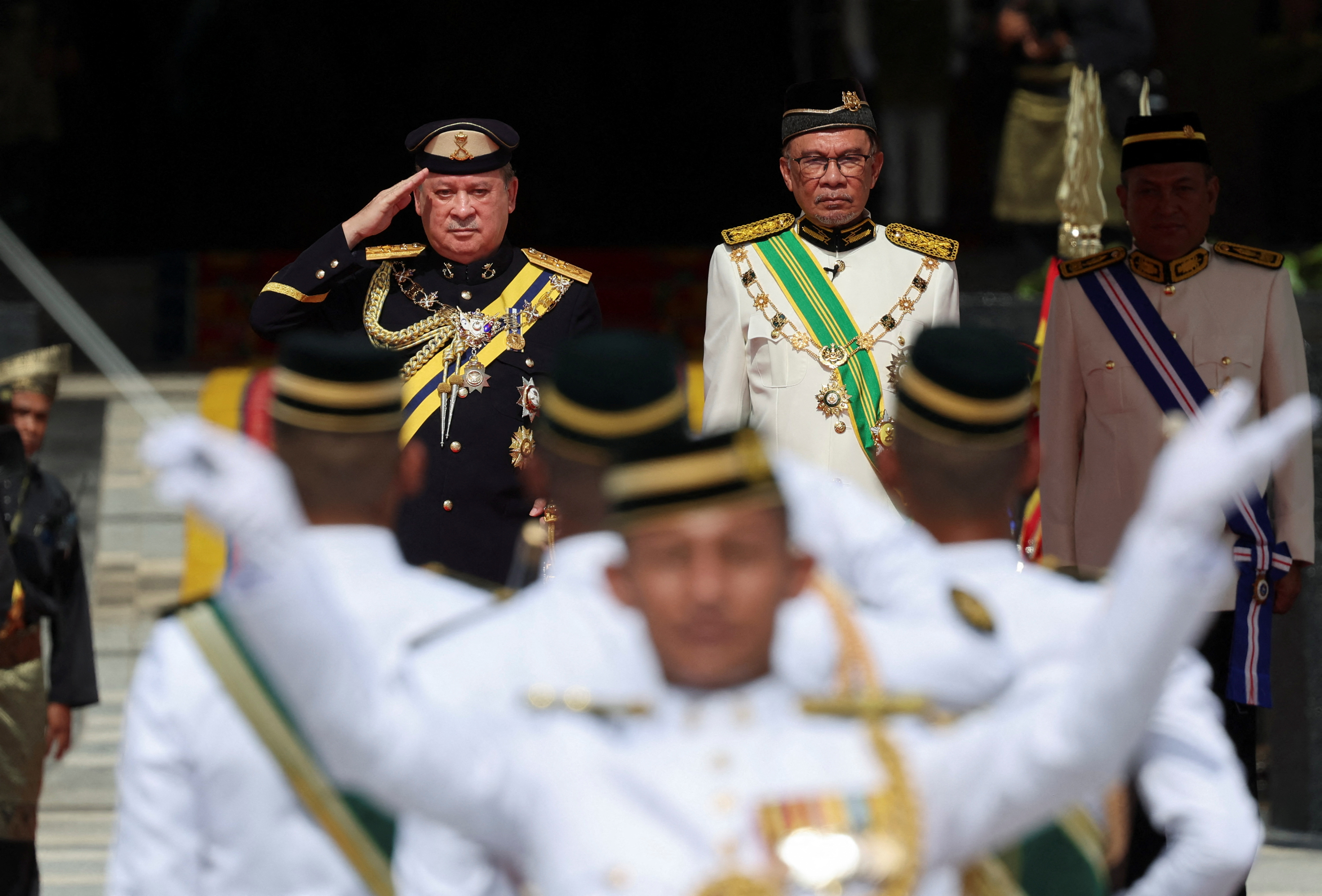 Sultan Ibrahim Sultan Iskandar salutes the guard of honour, beside Malaysia's Prime Minister Anwar Ibrahim at the National Palace. They are both in ceremonial uniforms/ Sultan Ibrahim's is black and Anwar's white with a green sash.