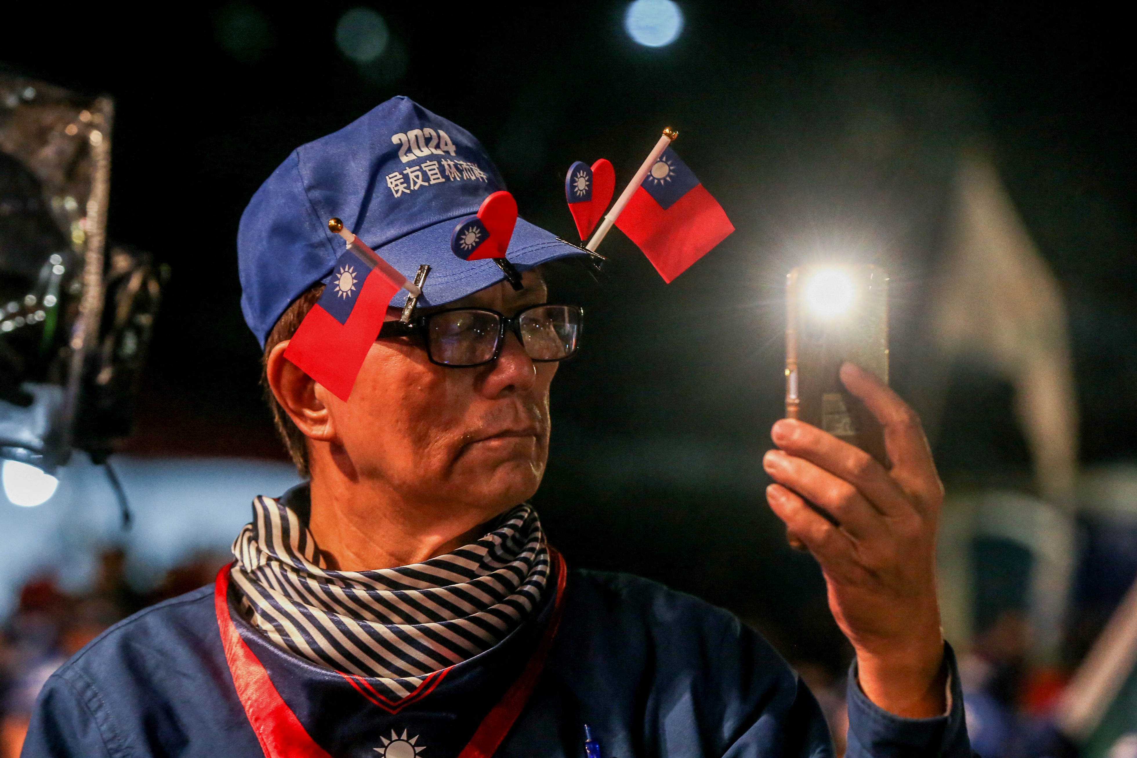 A KMT supporter at a rally. He is an older man and wearing a blue cap decorated with small Taiwan flags and hearts.