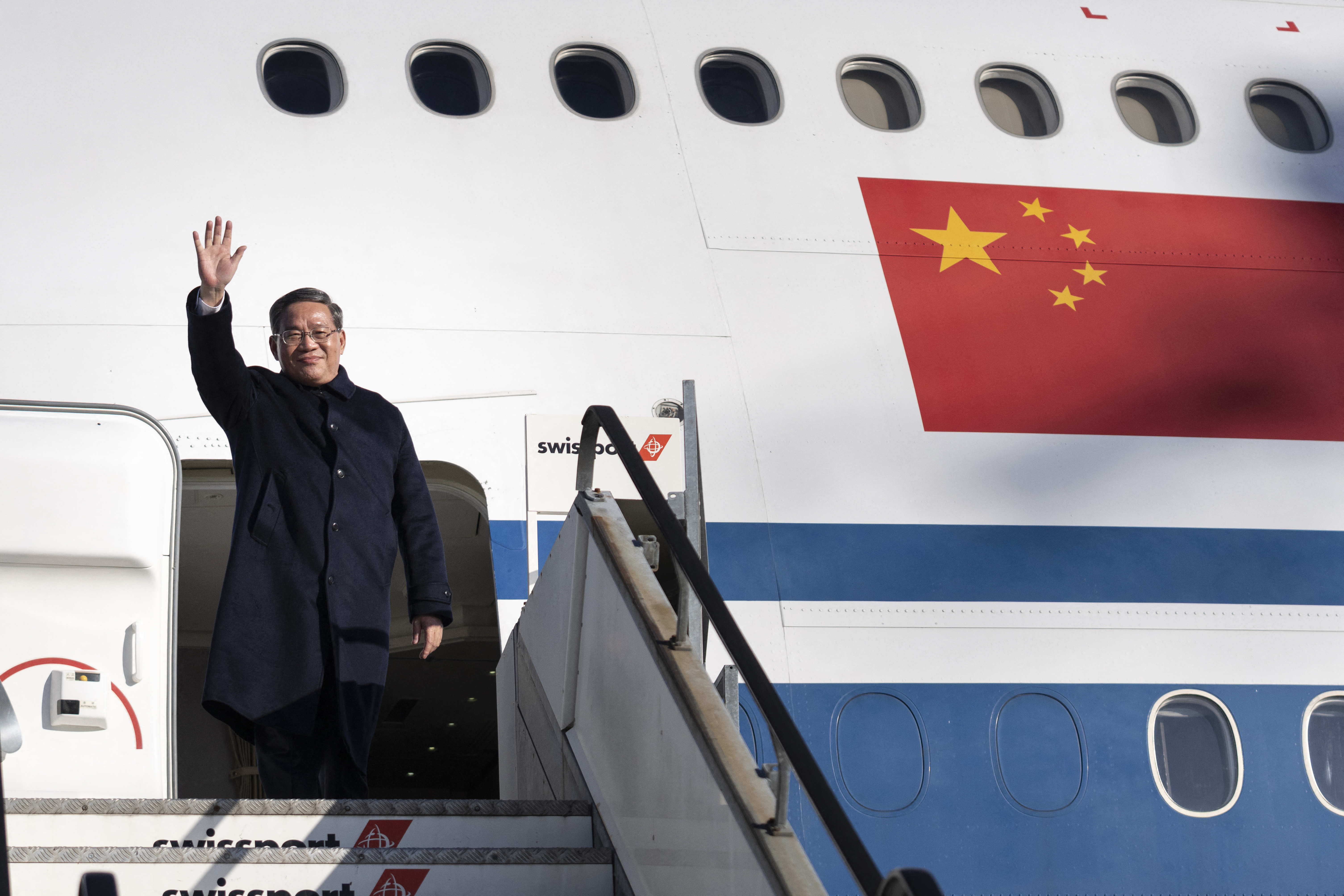Li Qiang at the top of the steps as he gets off an Air China plane in Zurich. He is wearing a black coat and waving.