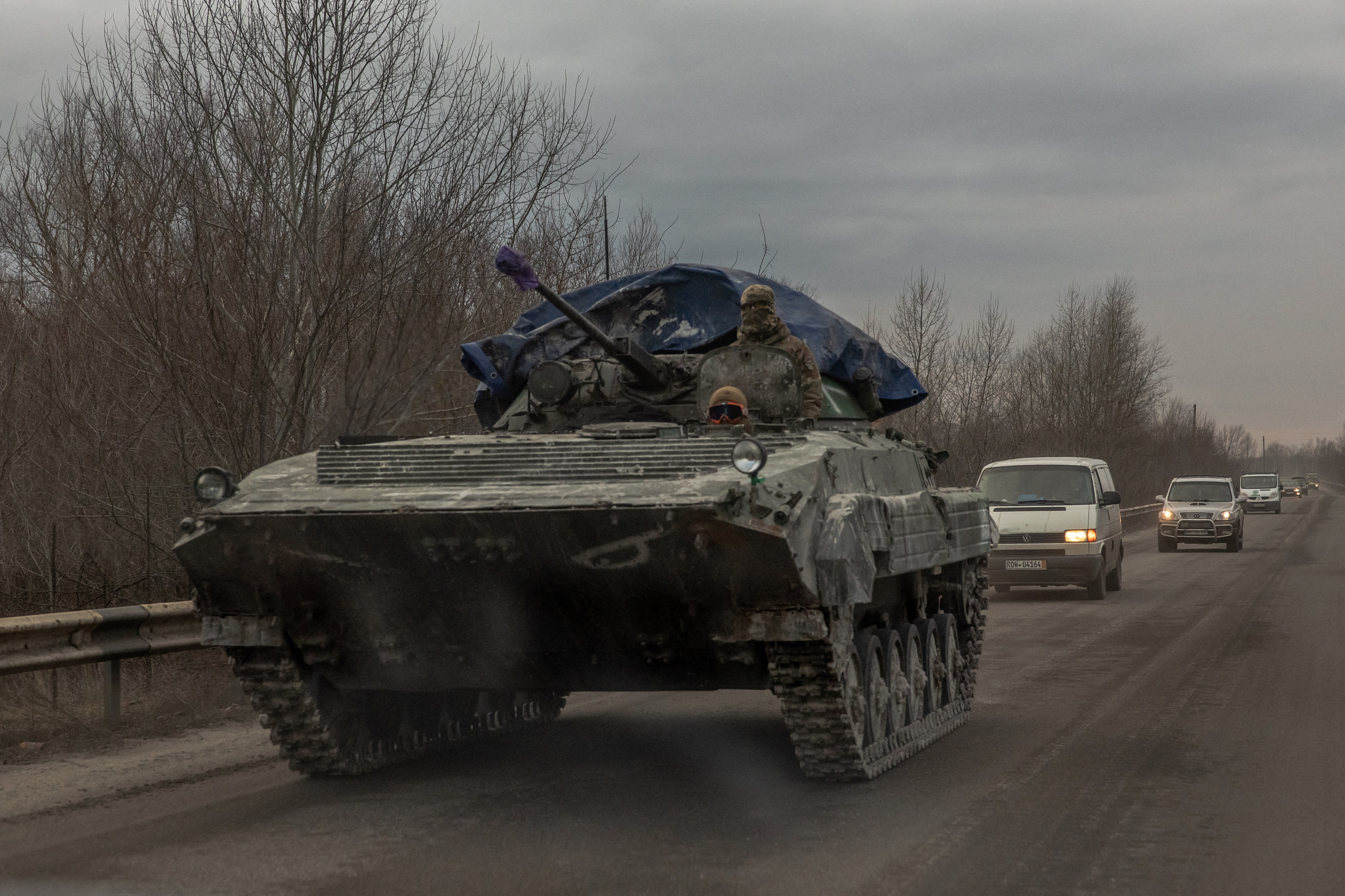 Ukrainian soldiers on top of an armoured vehicle on a road in Lyman. It looks grey and cold. There are cars on the road behind them.
