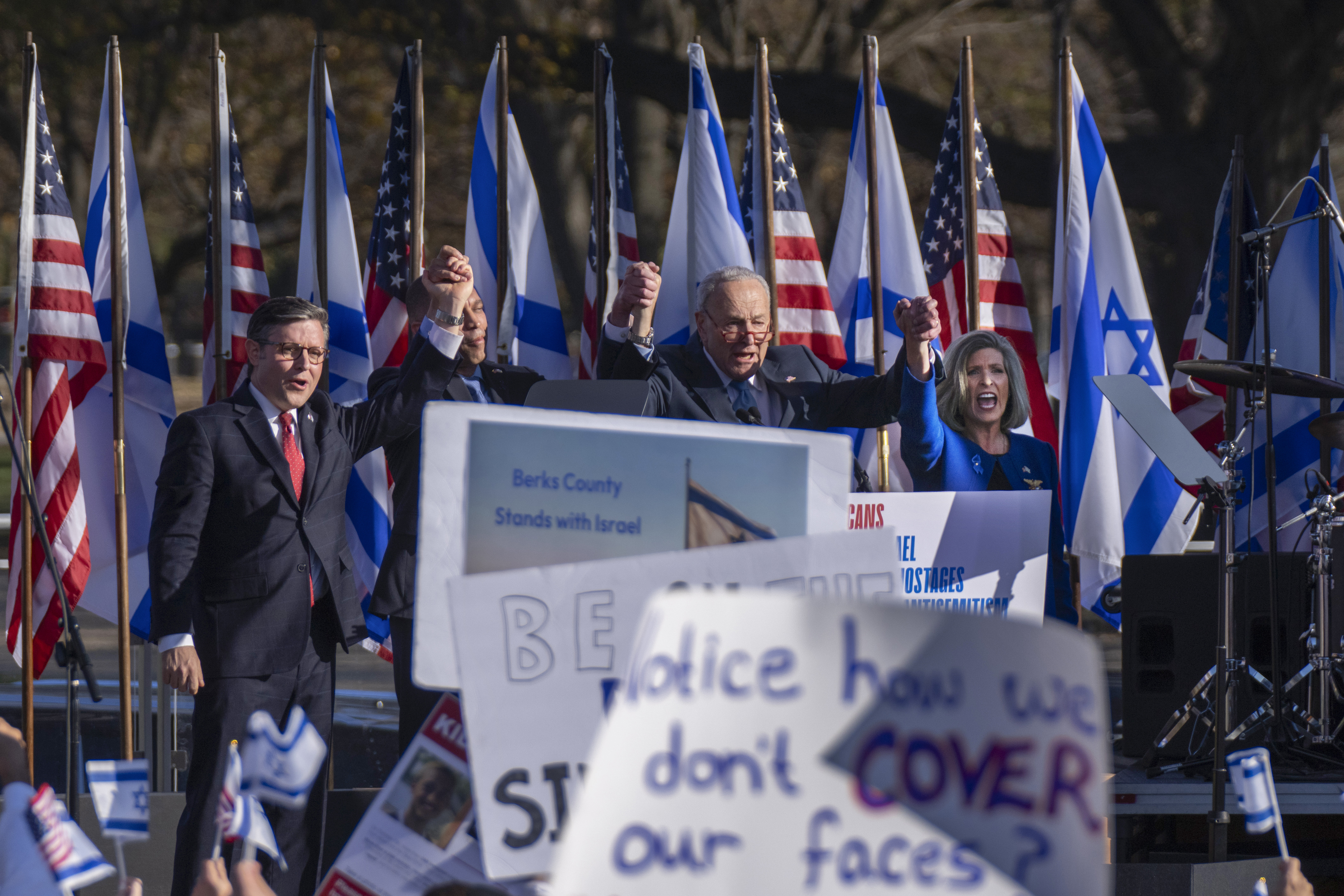 Protesters gather in Washington, DC, to see Chuck Schumer and other Congressional leaders speak at a March for Israel. The stage for the speakers is set up with US and Israeli flags, and audience members wave signs.