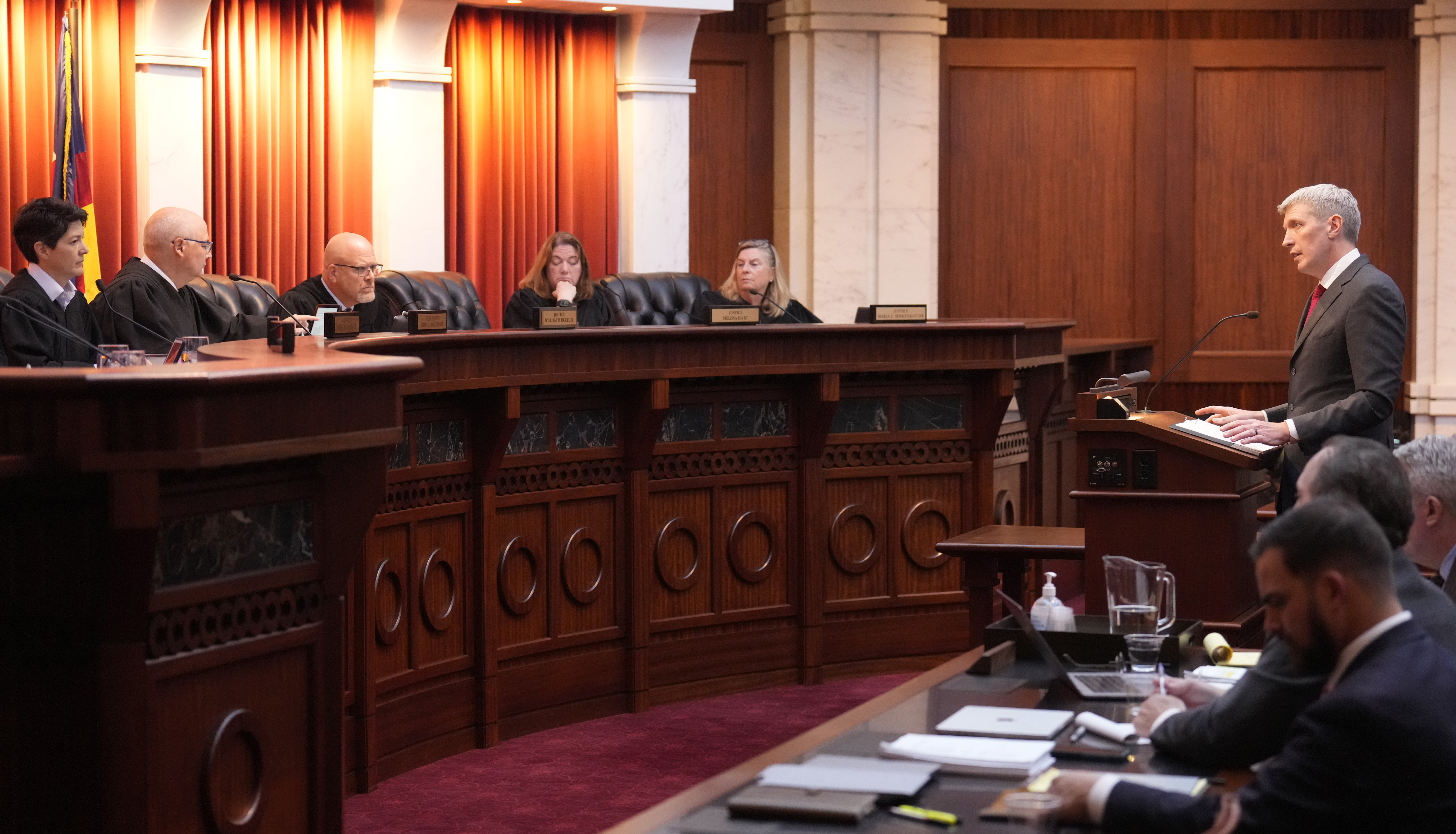 Attorney Eric Olson stands behind a podium in a wood-lined courtroom. Behind a raised panel table before him sit a row of judges.