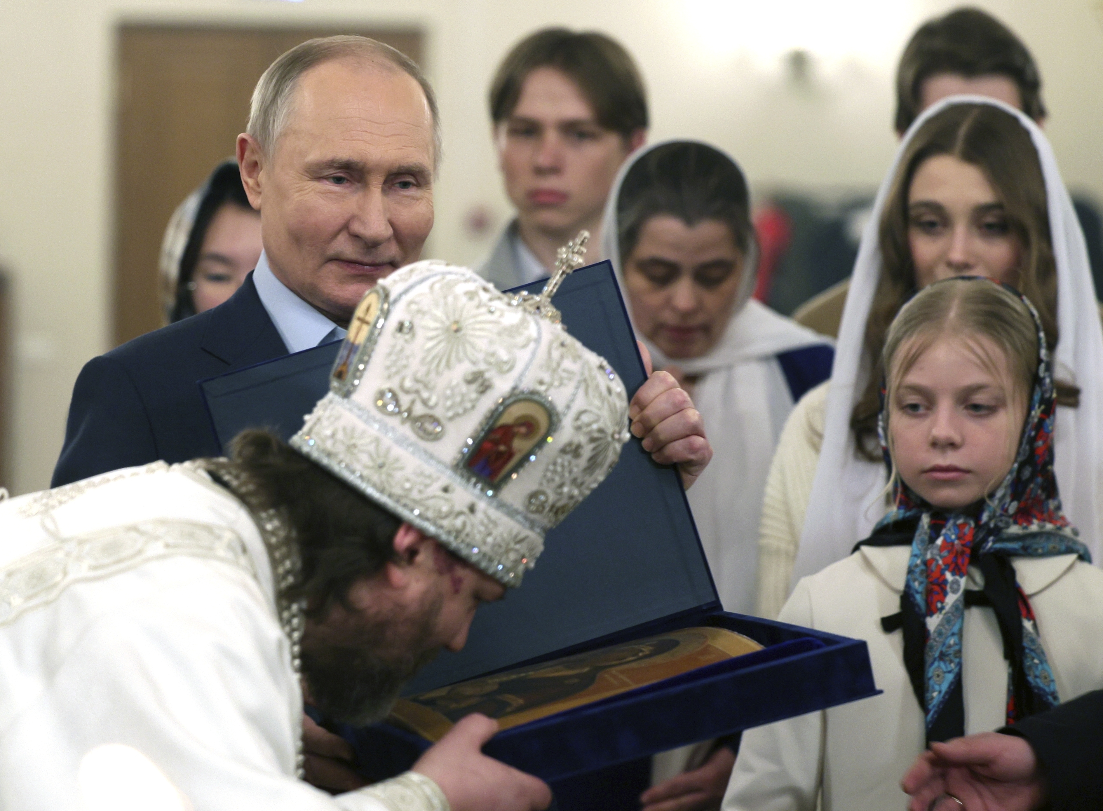 Putin smiling as he stands with the families of soldiers killed in Ukraine. A Russian Orthodox priest in white robes is kissing an icon in front of them.