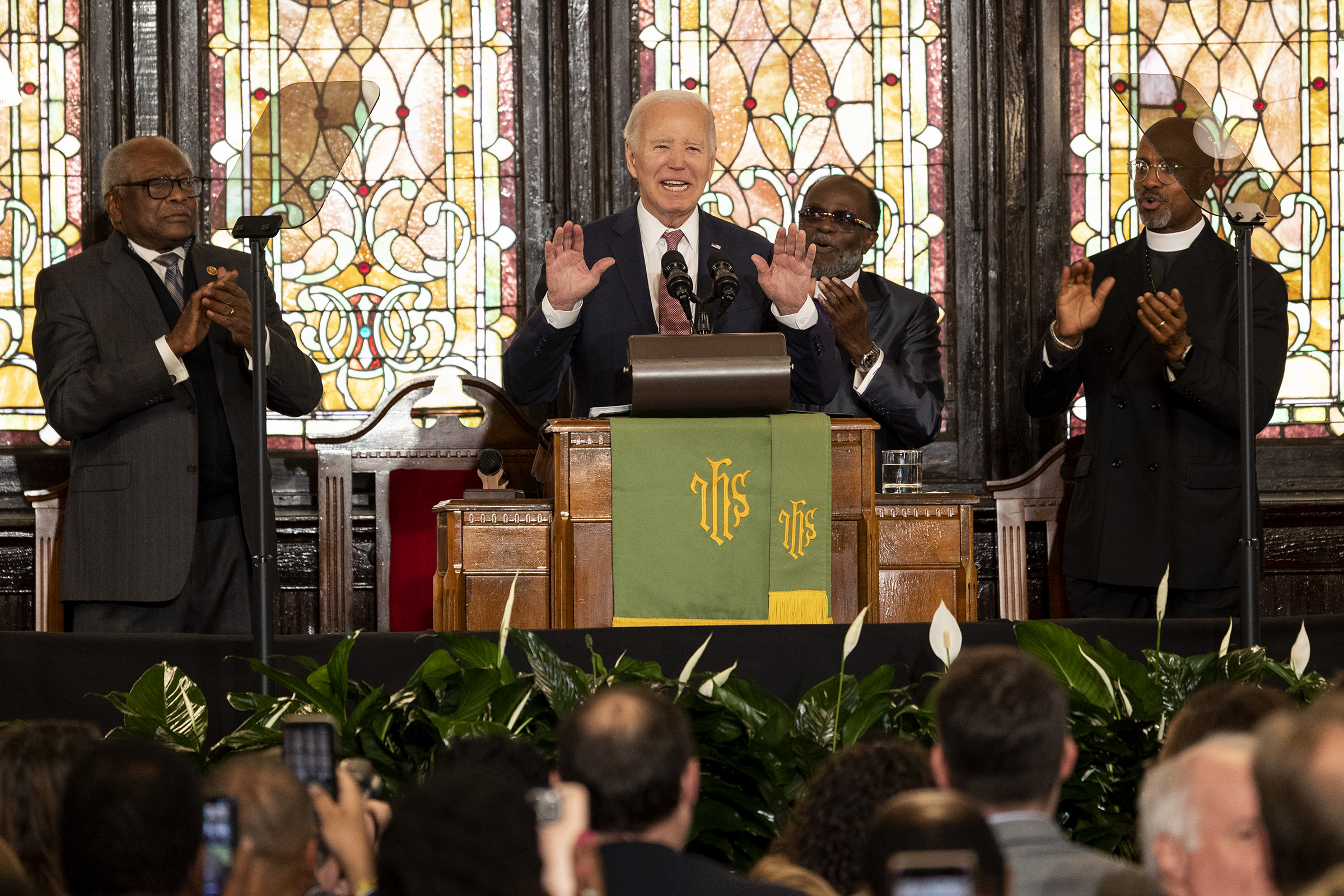 Joe Biden stands at the wooden pulpit of Mother Emanuel AME church in South Carolina, flanked by church leaders. Behind him are stained glass windows, and calla lilies and audience members sit in front of him.