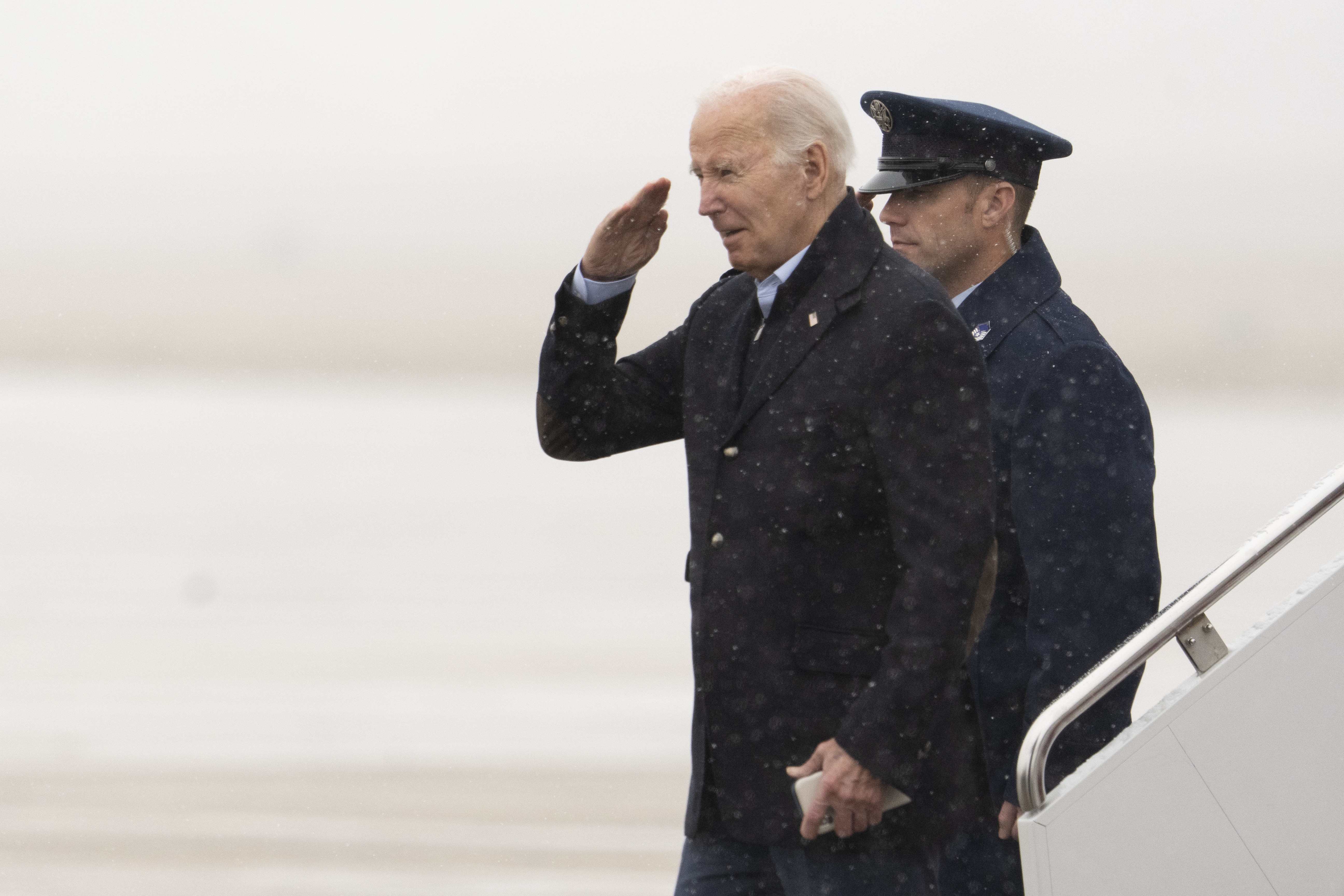 Joe Biden descends the steps of Air Force One dressed in a long, dark winter coat. He salutes as he walks. Behind him is a military service member or some other member of his security team.