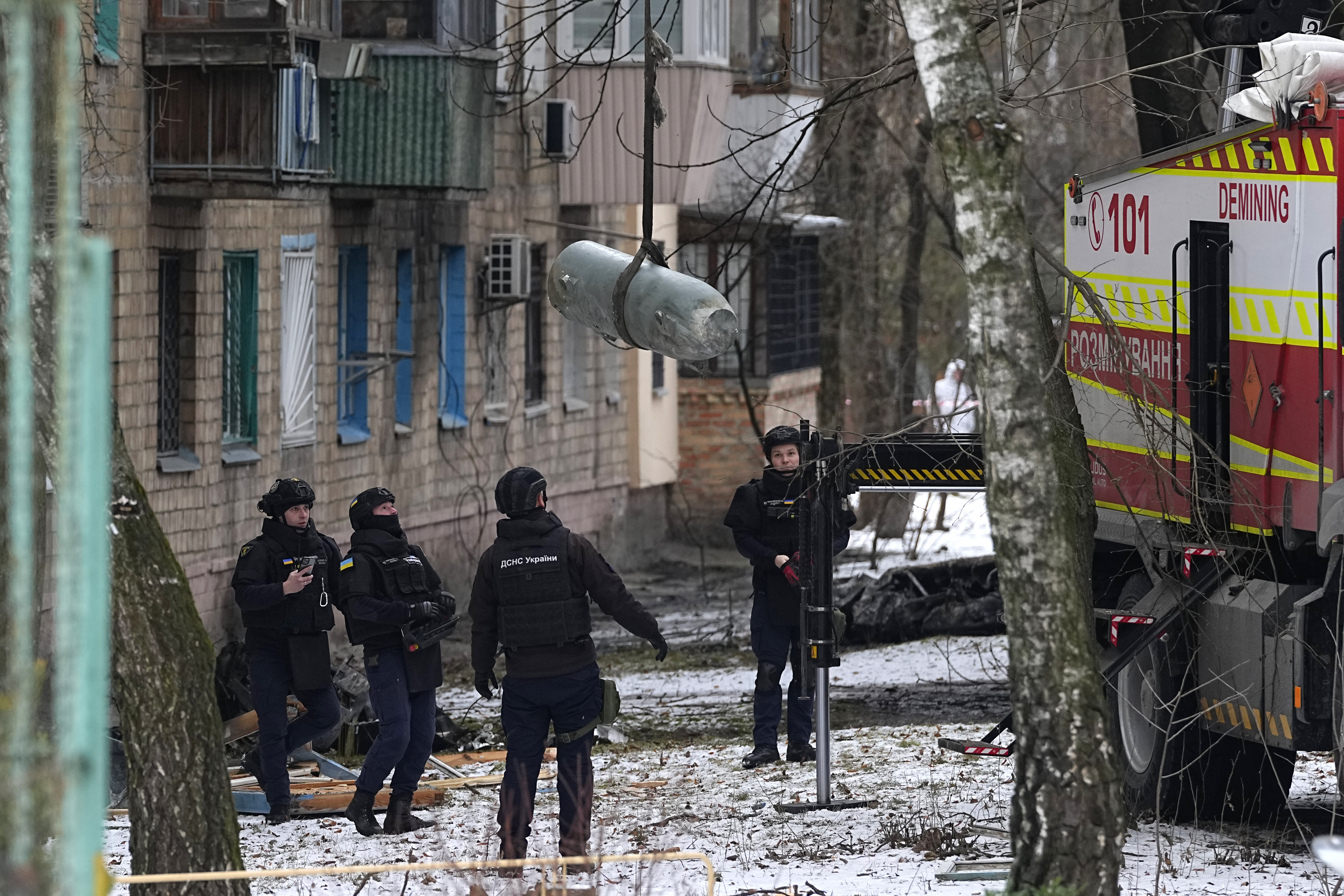 Sappers load an unexploded missile into a truck, The missile is being lifted into the air. The men are standing next to a damaged apartment block. There's snow on the ground.