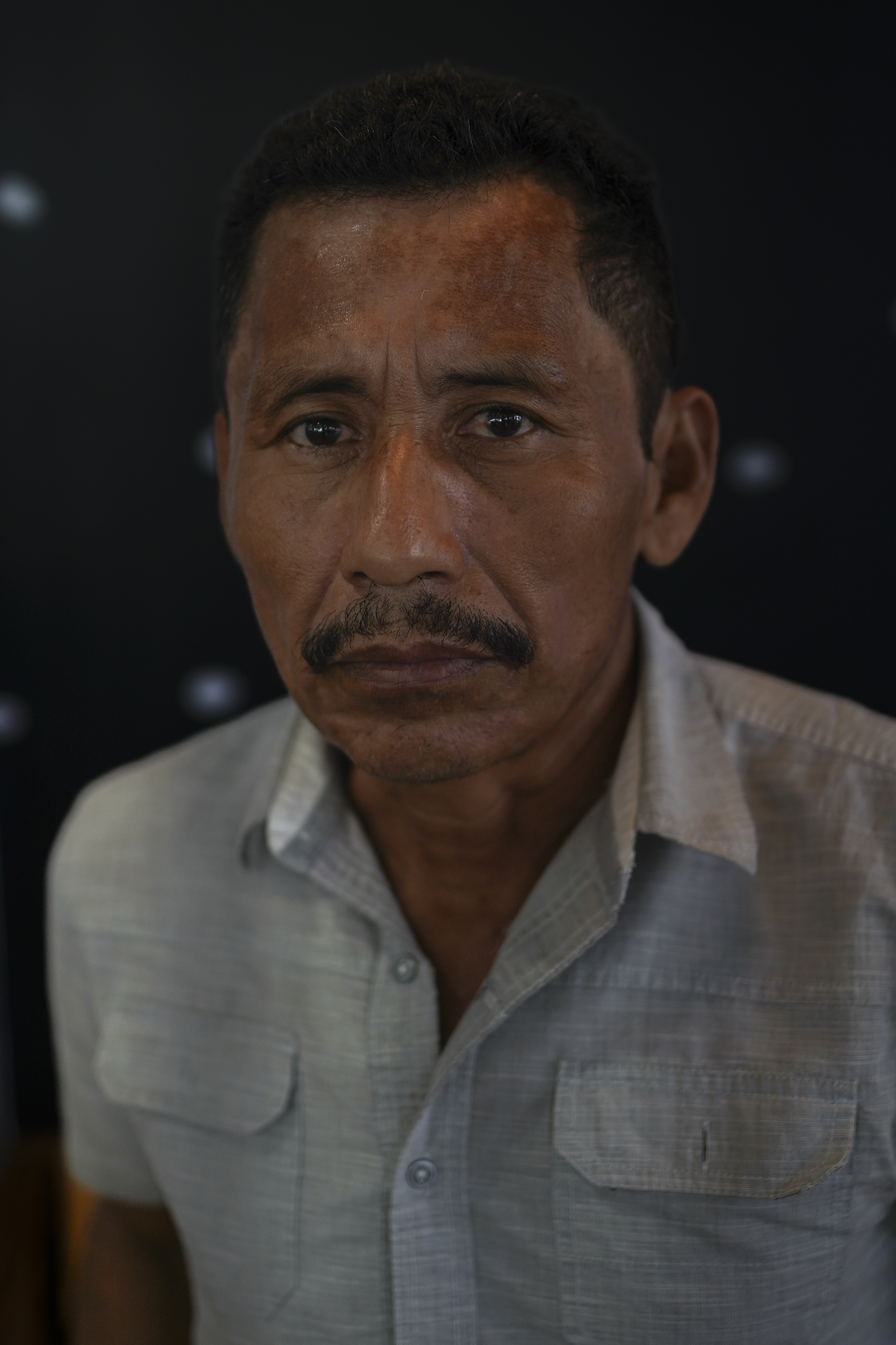 A middle-aged man with a mustache and a collared shirt stares into the camera.