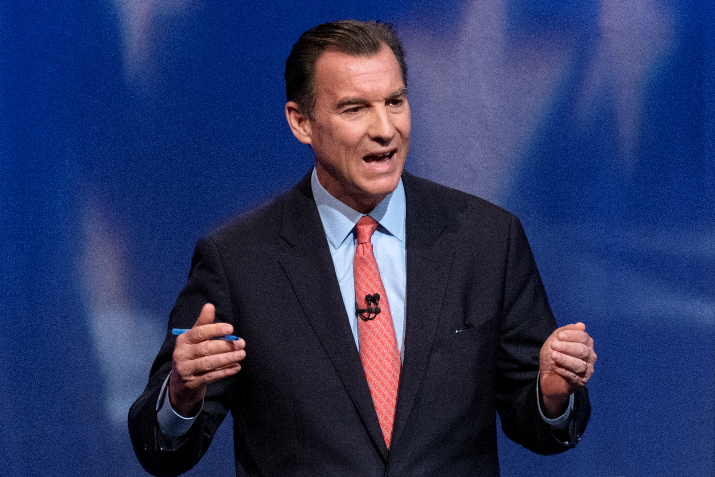Tom Suozzi, dressed in a dark suit and red tie, speaks with his hands raised by his side in gesture.