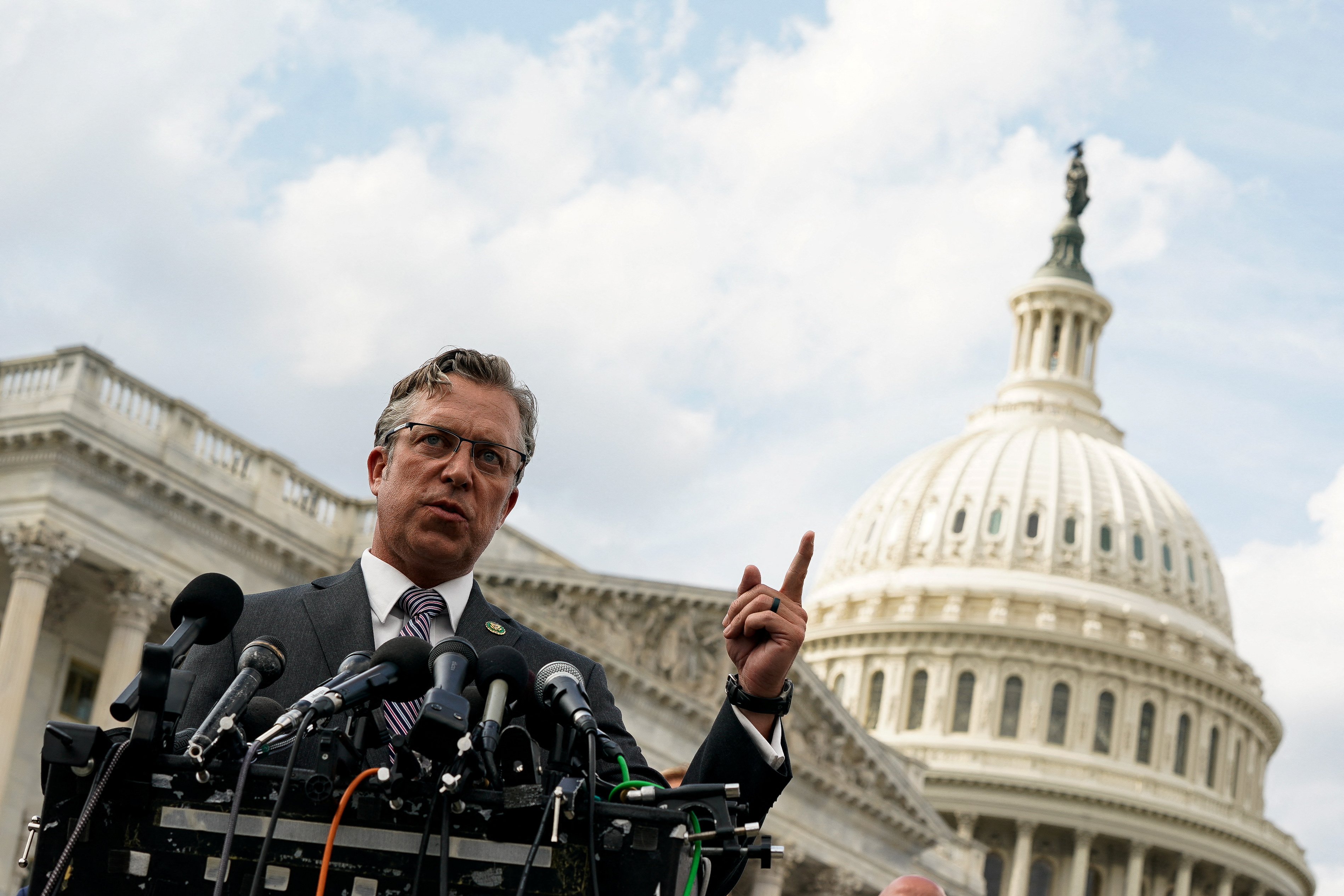 Andy Ogles stands behind a microphone-filled podium and lifts a finger in gesture. The dome of the US Congress is visible behind him.