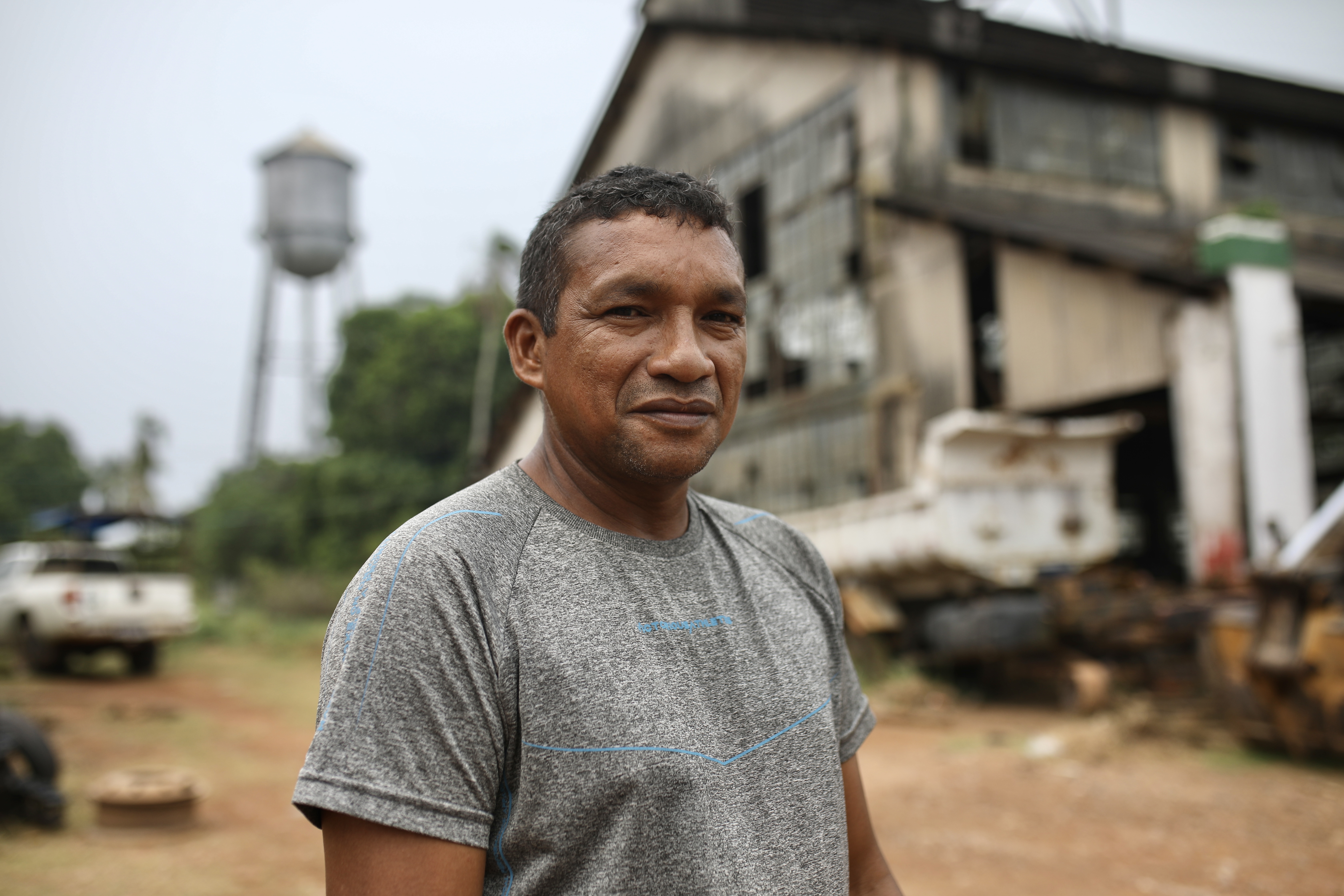 A man in a grey T-shirt stands in front of the abandoned warehouses at Fordlandia, an industrial town in the Amazon. A water tower is also visible behind him.