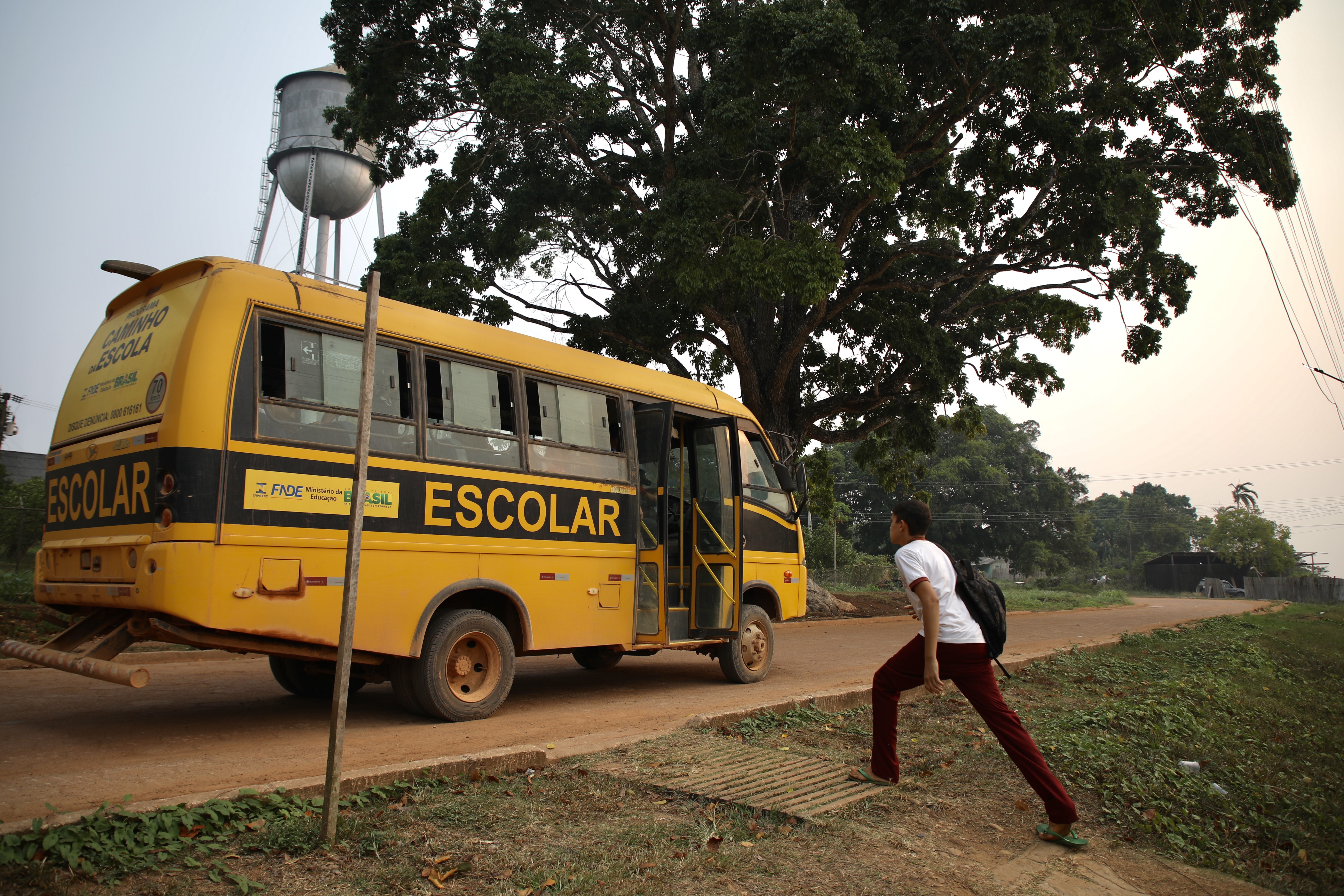 A young student takes wide strides to reach an orange school bus waiting for him on a dirt road.