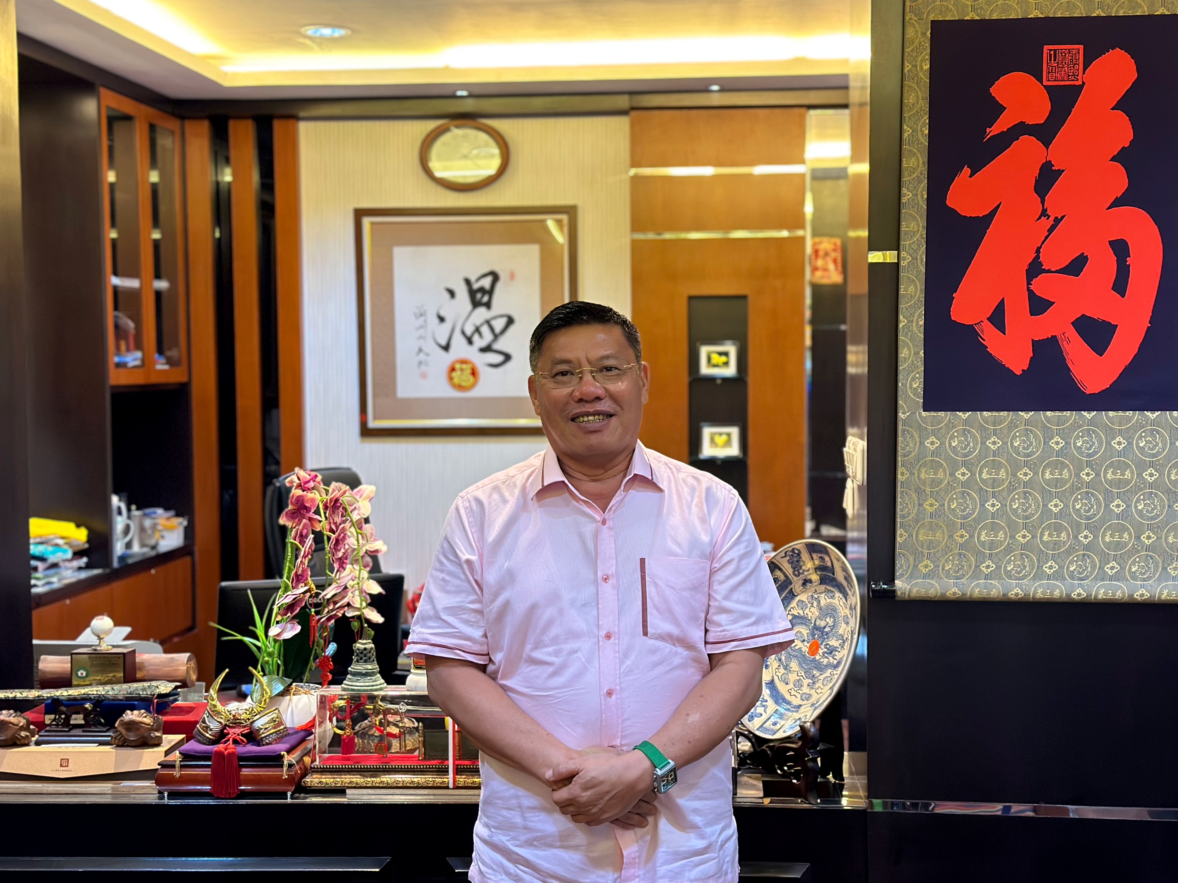 Fuidy Luckman in his office. He is standing in front of his desk. He's wearing a pink shirt and looks relaxed. 