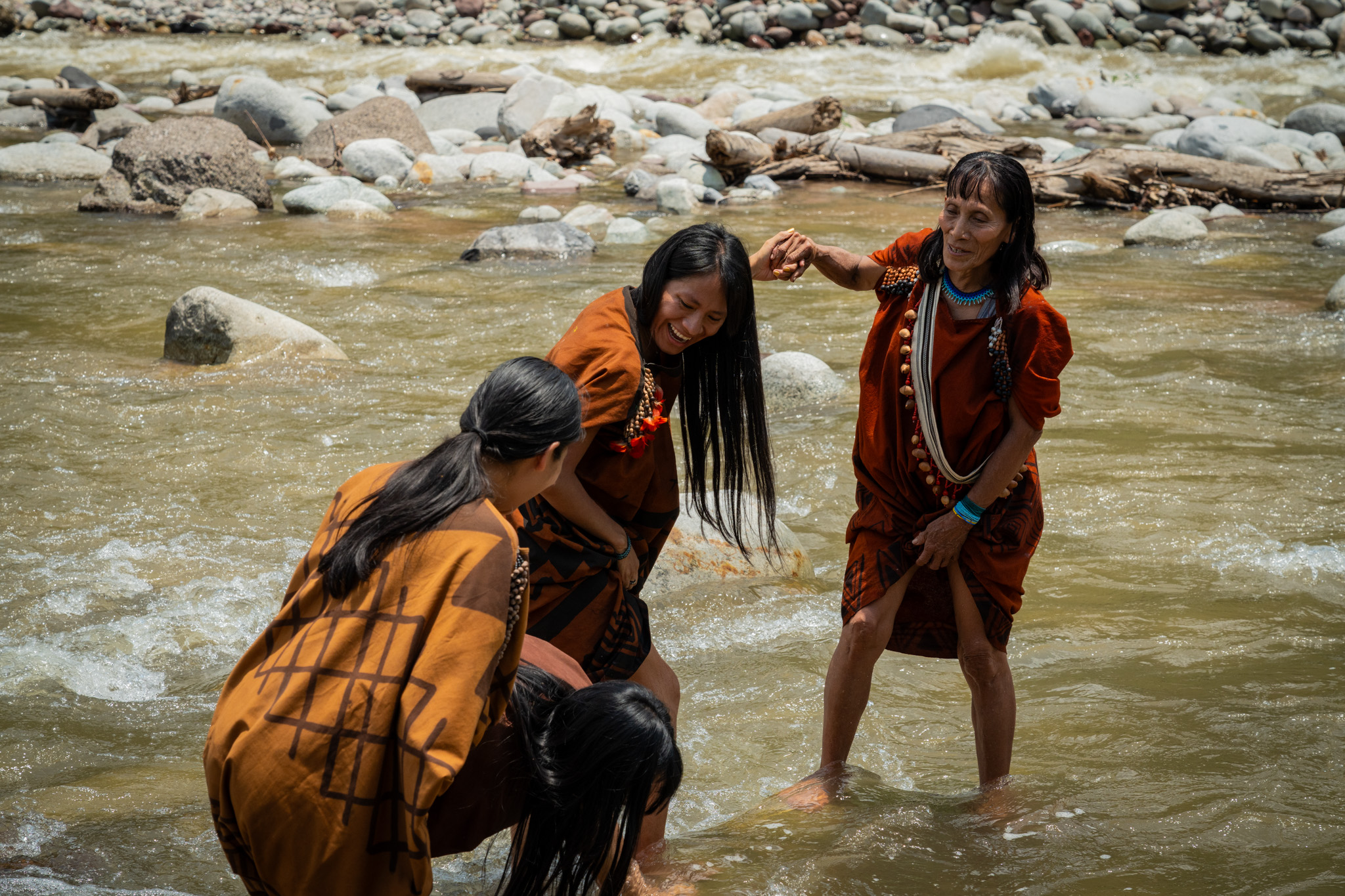 Three Indigenous Ashaninka women stand and smile in a river lined with rocks.